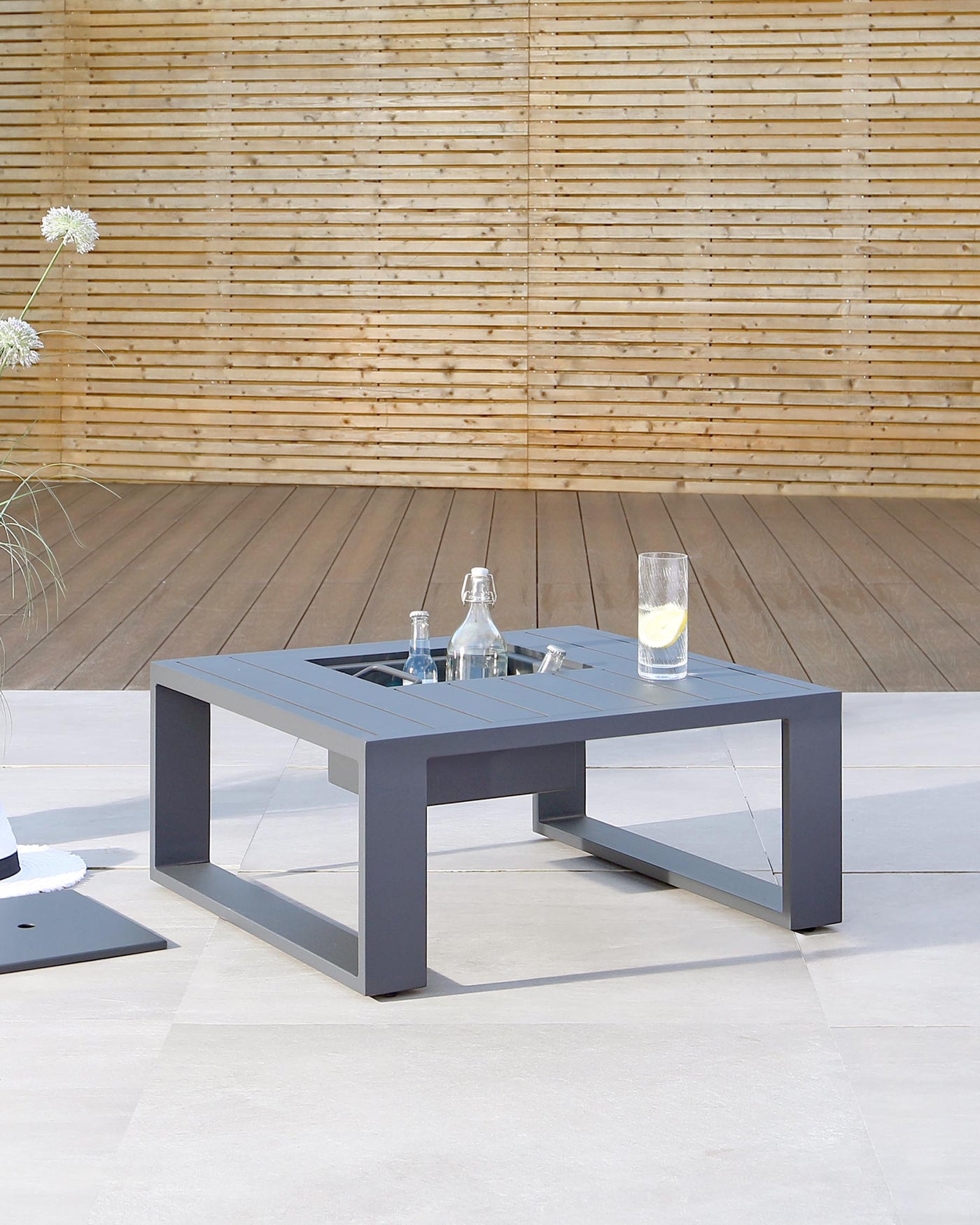Contemporary dark grey square outdoor coffee table with integrated ice bucket feature in the centre, showcased on a patio with light flooring and a bamboo backdrop.