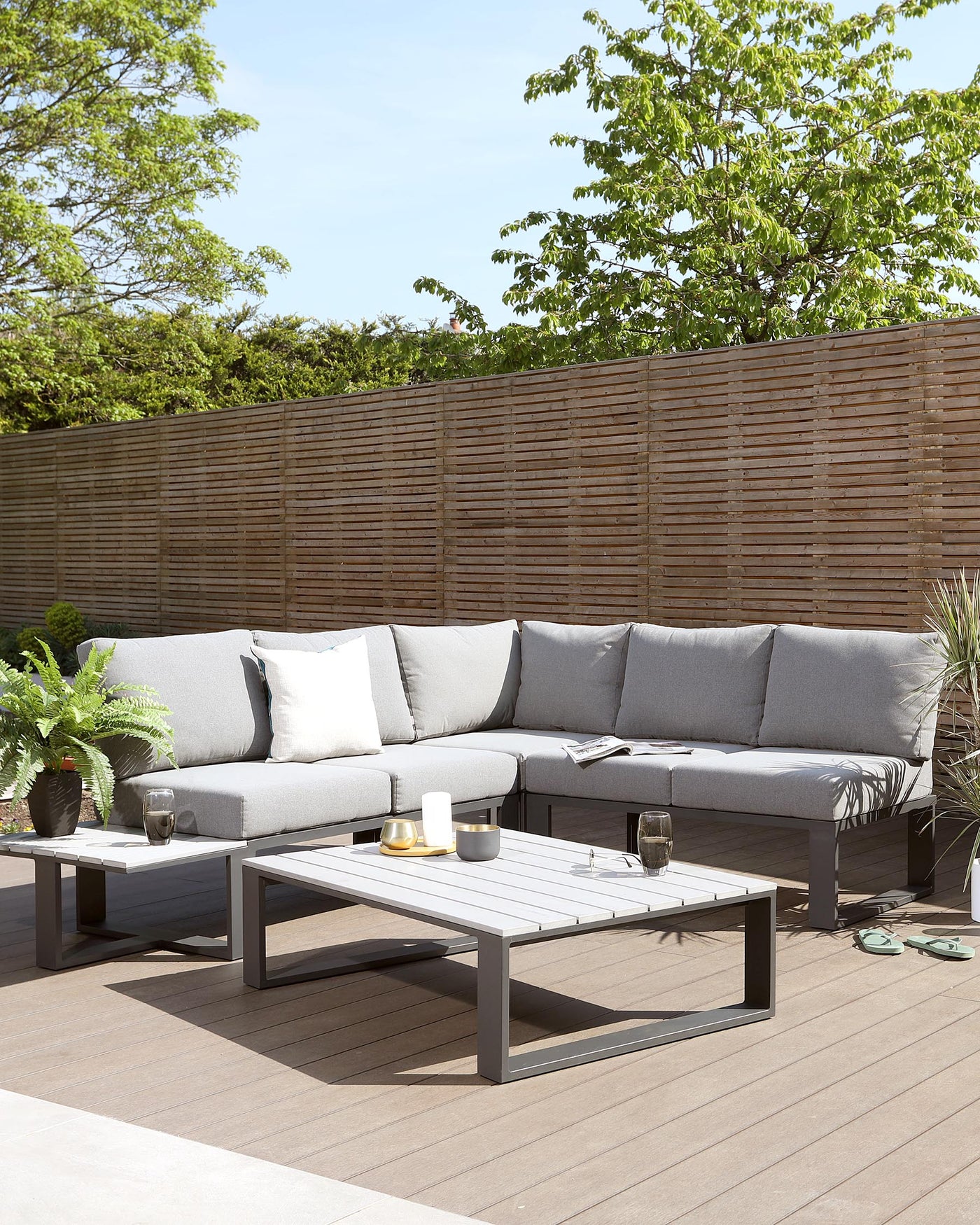 Outdoor patio furniture consisting of an L-shaped sectional sofa with light grey cushions and a matching low-profile rectangular coffee table in a grey finish, set on a wooden deck with decorative plants and a slatted privacy screen in the background.