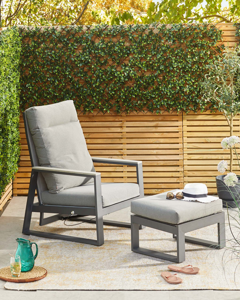 Contemporary outdoor furniture set featuring a grey metal frame recliner with matching cushions and a square metal ottoman with a cushion top, arranged on a pale patterned area rug. A green glass pitcher and drinking glass set upon a round woven tray rest near the ottoman.