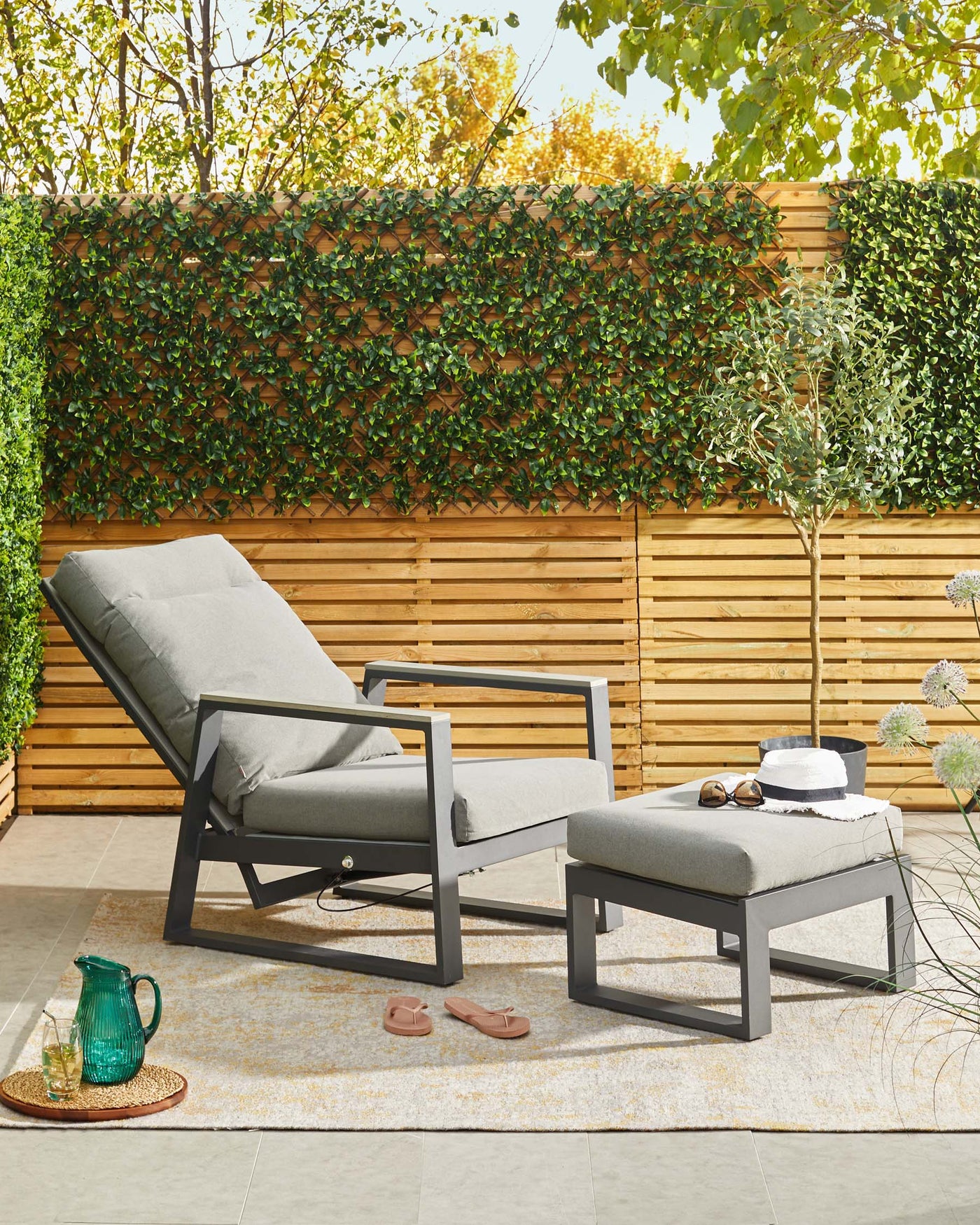 Outdoor lounge chair with a matching ottoman, both featuring a modern design with grey cushions and matte black frames, set against a natural backdrop with a wooden fence and greenery.