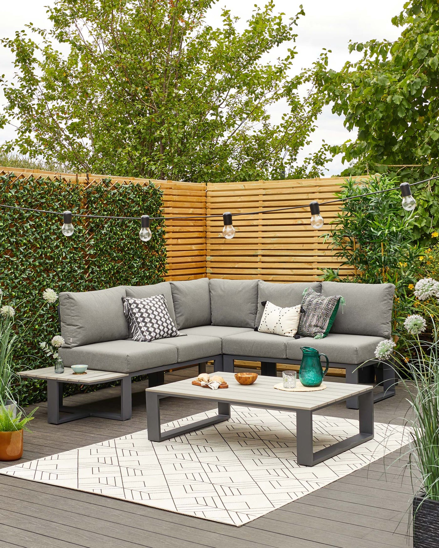 Outdoor sectional sofa with grey cushions on a dark frame situated on a wooden deck, accompanied by a matching low rectangular coffee table also in dark tones. The setup is completed with decorative pillows, a textured outdoor rug with geometric patterns, and ambient string lights overhead. A few potted plants add a touch of greenery to the cosy setup.