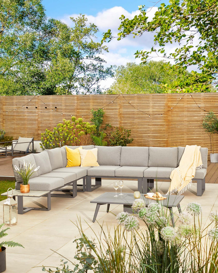 Modern outdoor sectional sofa in grey with a matching low coffee table, featuring light grey cushions, yellow accent pillows, and a cream-colored throw blanket. The furniture is set on a patio surrounded by lush greenery and a wooden fence.