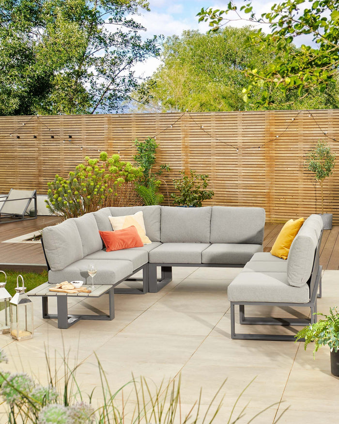 Contemporary outdoor sectional sofa with light grey cushions on a minimalistic metal frame, paired with a matching ottoman and a square low-profile metal coffee table. The furniture is accessorized with coral and yellow accent pillows, set against a backdrop of a manicured garden with wooden fencing.