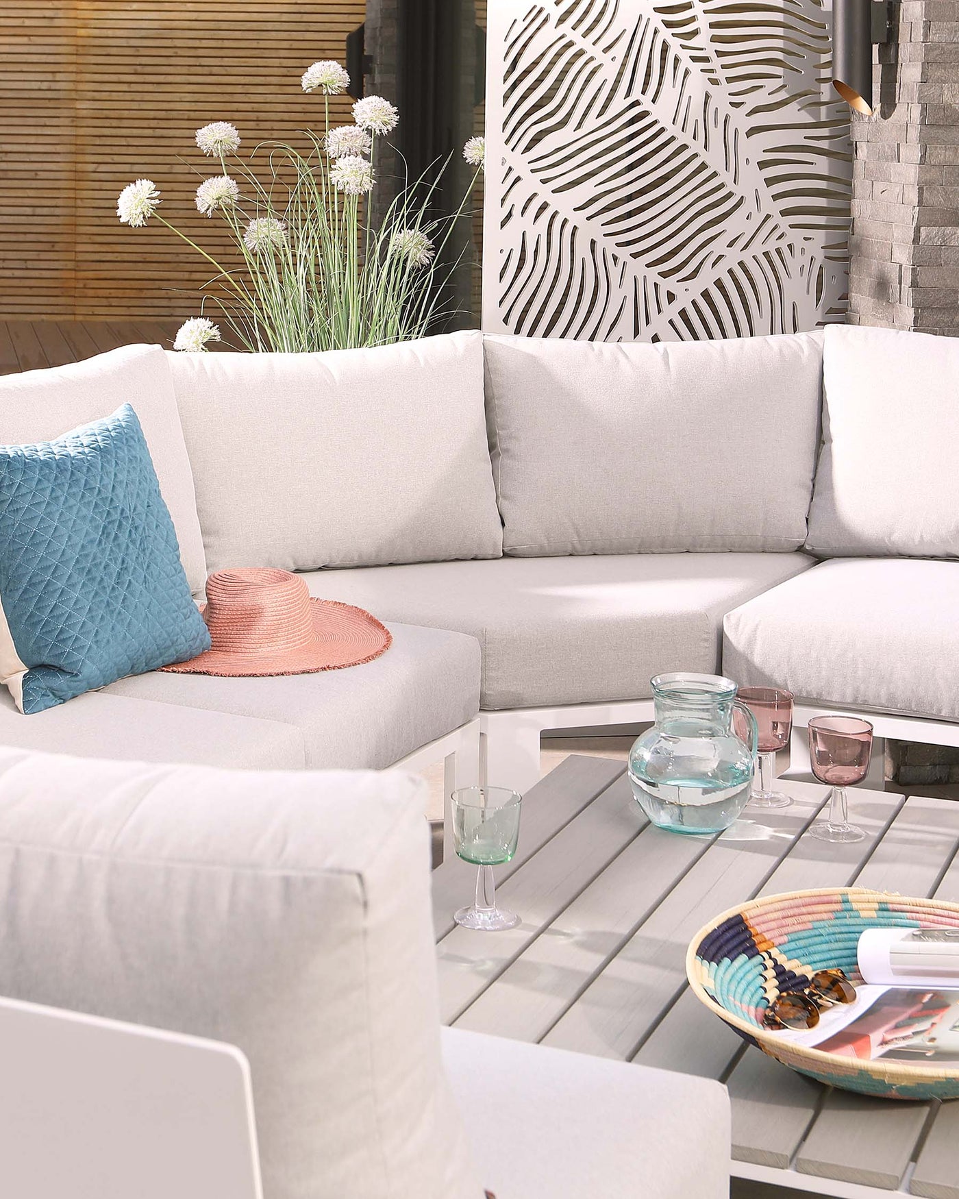Modern outdoor corner sectional sofa in light grey with clean lines and plush cushions accompanied by a low-profile, slatted coffee table in a coordinating grey tone. Decorative pillows in varying hues of blue and pink add a pop of colour.