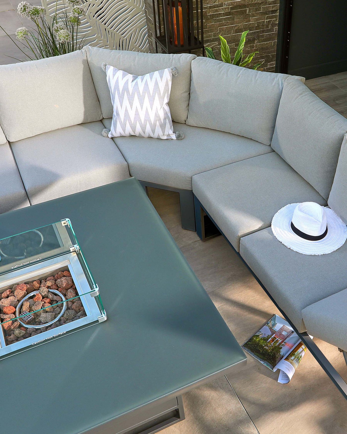 A modern outdoor corner sectional sofa in a light grey fabric with clean lines and minimalistic style, complemented by a matching low-profile coffee table with a frosted glass display compartment filled with decorative stones. The set is accessorized with a patterned throw pillow, a casual white sun hat, and a lifestyle magazine, arranged on a balcony with potted plants and a textured wall as a backdrop.
