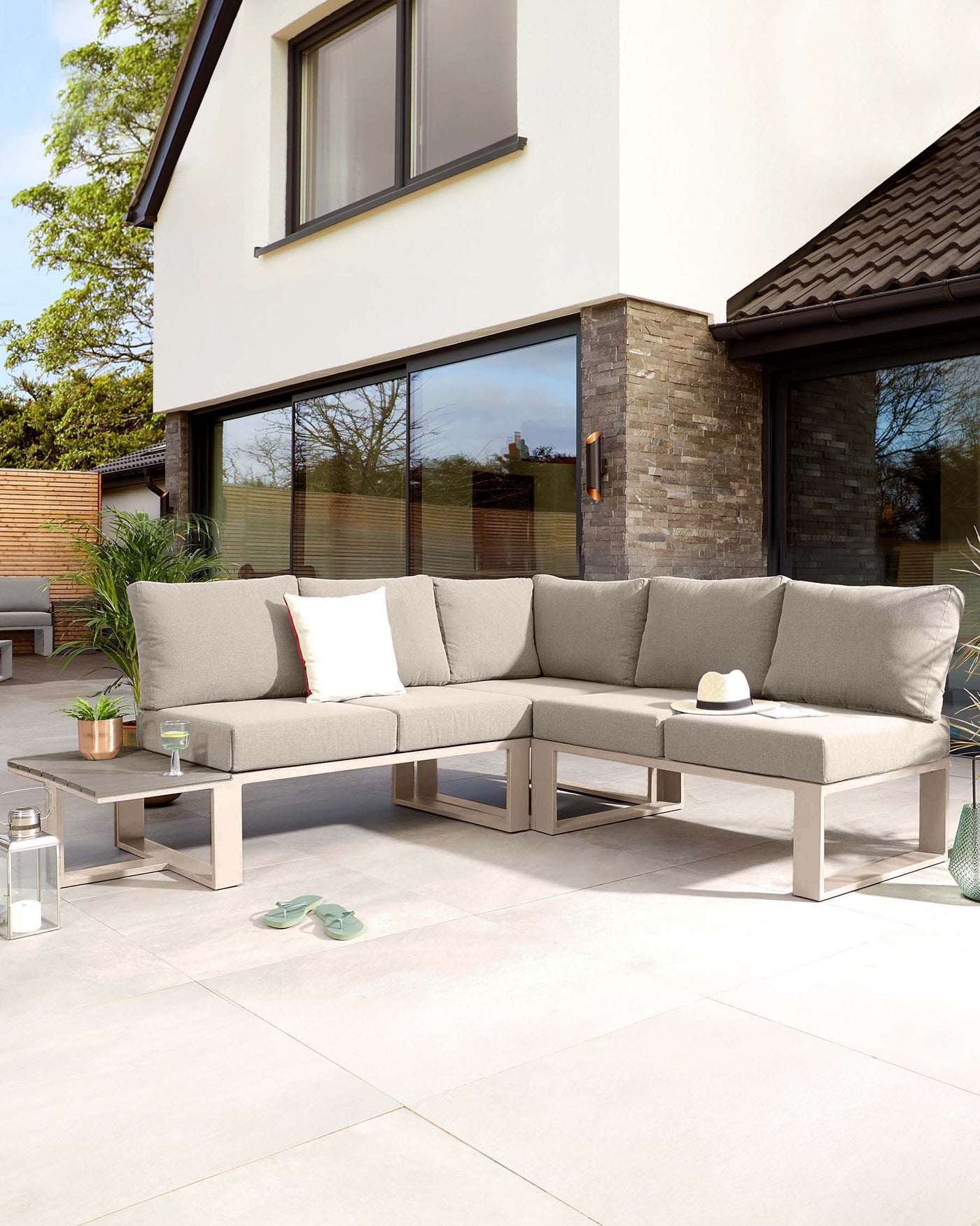 Contemporary outdoor L-shaped sectional sofa with beige cushions on a lightweight, minimalist aluminium frame, accompanied by a rectangular low-height coffee table featuring a matching aluminium base and a light-coloured tabletop, displayed on a patio setting.