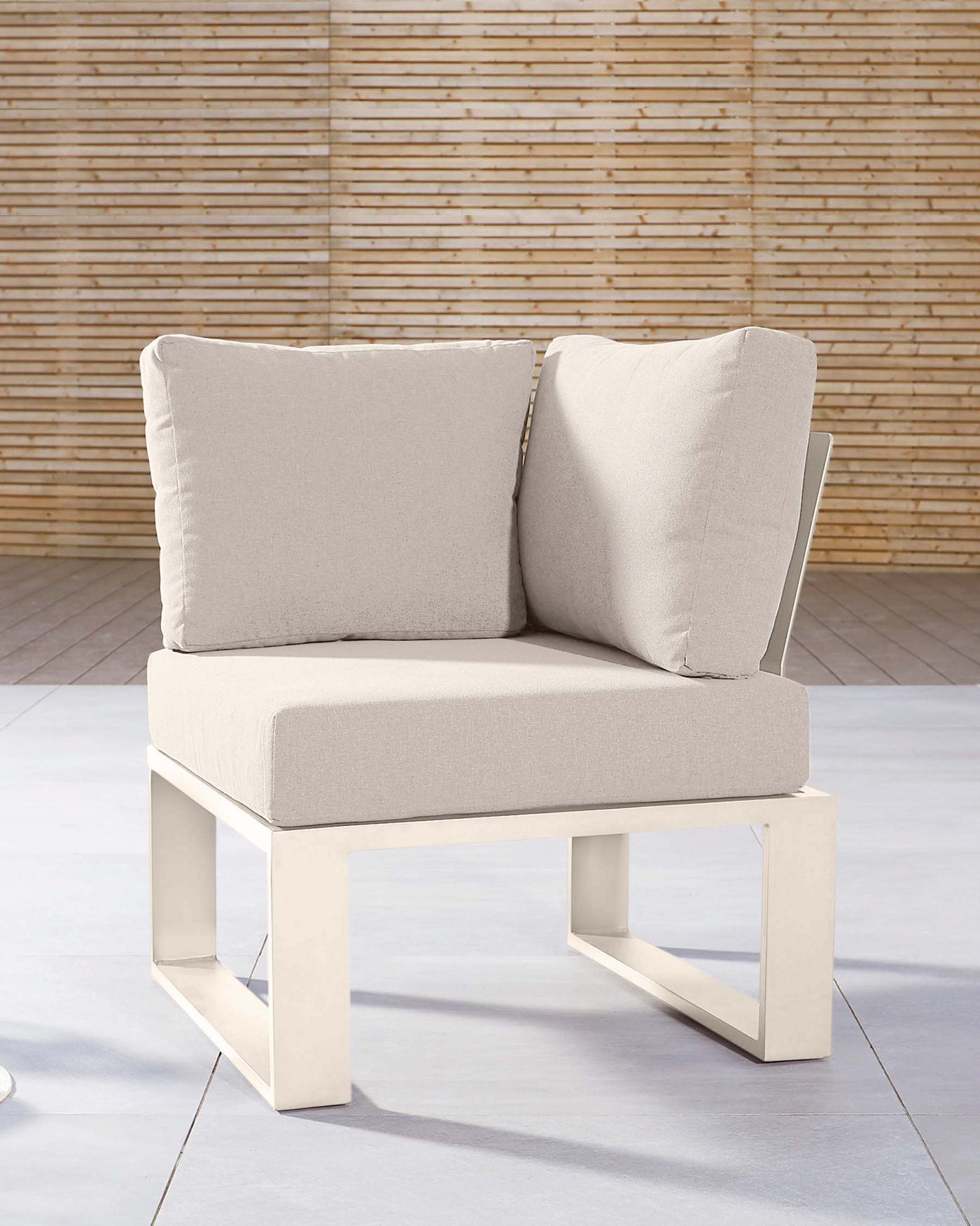 Modern single-seater sofa with a minimalist design, featuring a solid base with a clean-lined rectangular silhouette, upholstered in a light, neutral fabric. The seat is complemented by plush back and side cushions, providing comfort and support. The simple yet stylish piece is set against a bamboo backdrop, suggesting an outdoor or sunlight-filled indoor setting.