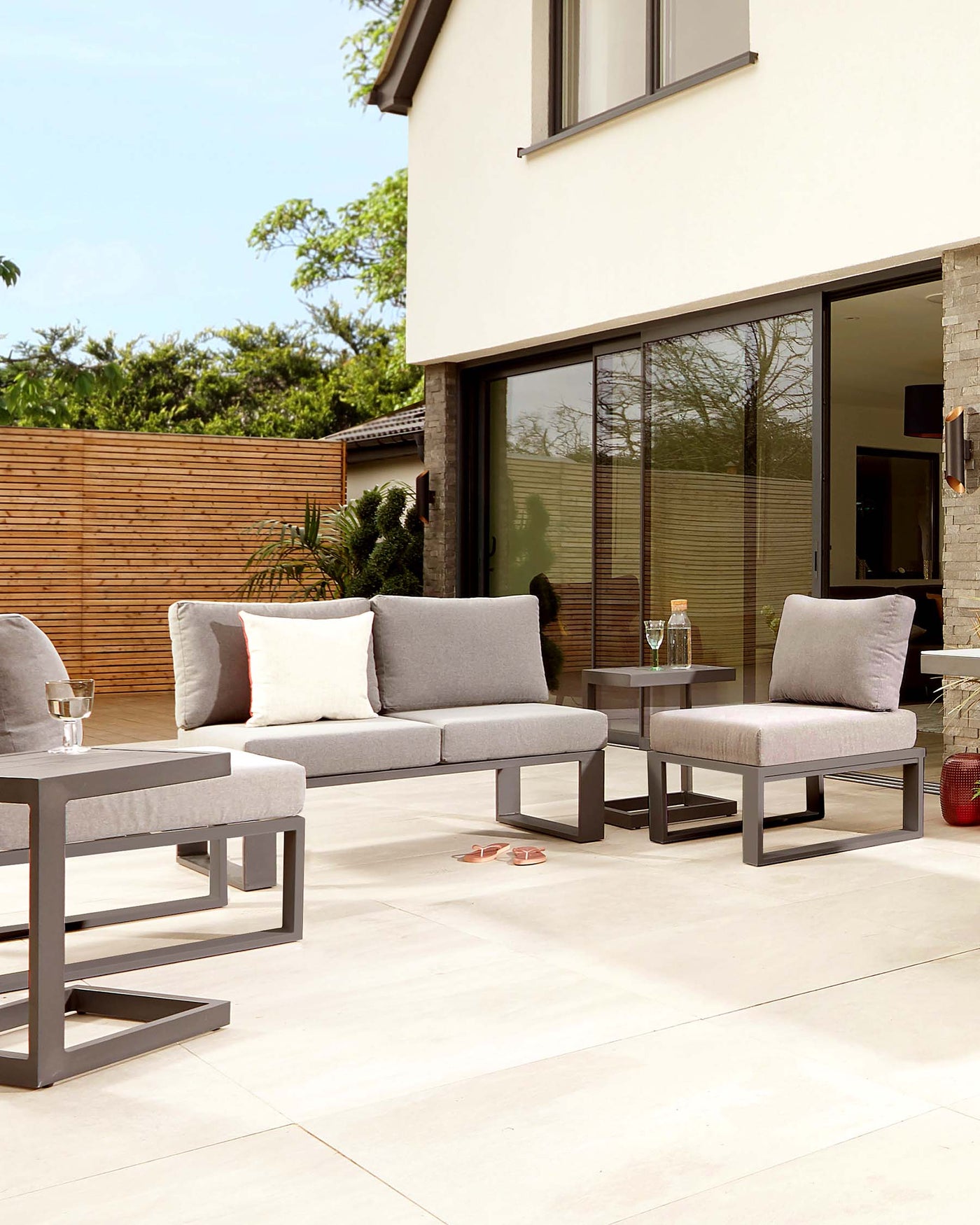 Modern outdoor furniture consisting of two grey upholstered single-seat lounge chairs with backrests, one double-seat lounge chair with a matching backrest, and a rectangular low-profile coffee table with a flat grey surface, all with a sleek dark grey metal frame. The set is arranged on a beige tiled patio, and decorative cushions and a carafe with glasses complete the scene.