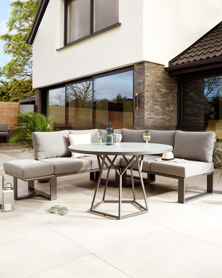 Modern outdoor furniture set featuring a geometric metal-framed round table with a glass top, and a sectional corner sofa with padded seats and backrests in a matching grey tone. The set includes a separate square ottoman that can double as seating or a footrest. The furniture is displayed on a patio, complemented by decorative cushions, a wine glass, and a lantern, creating an inviting al fresco dining and lounging area.