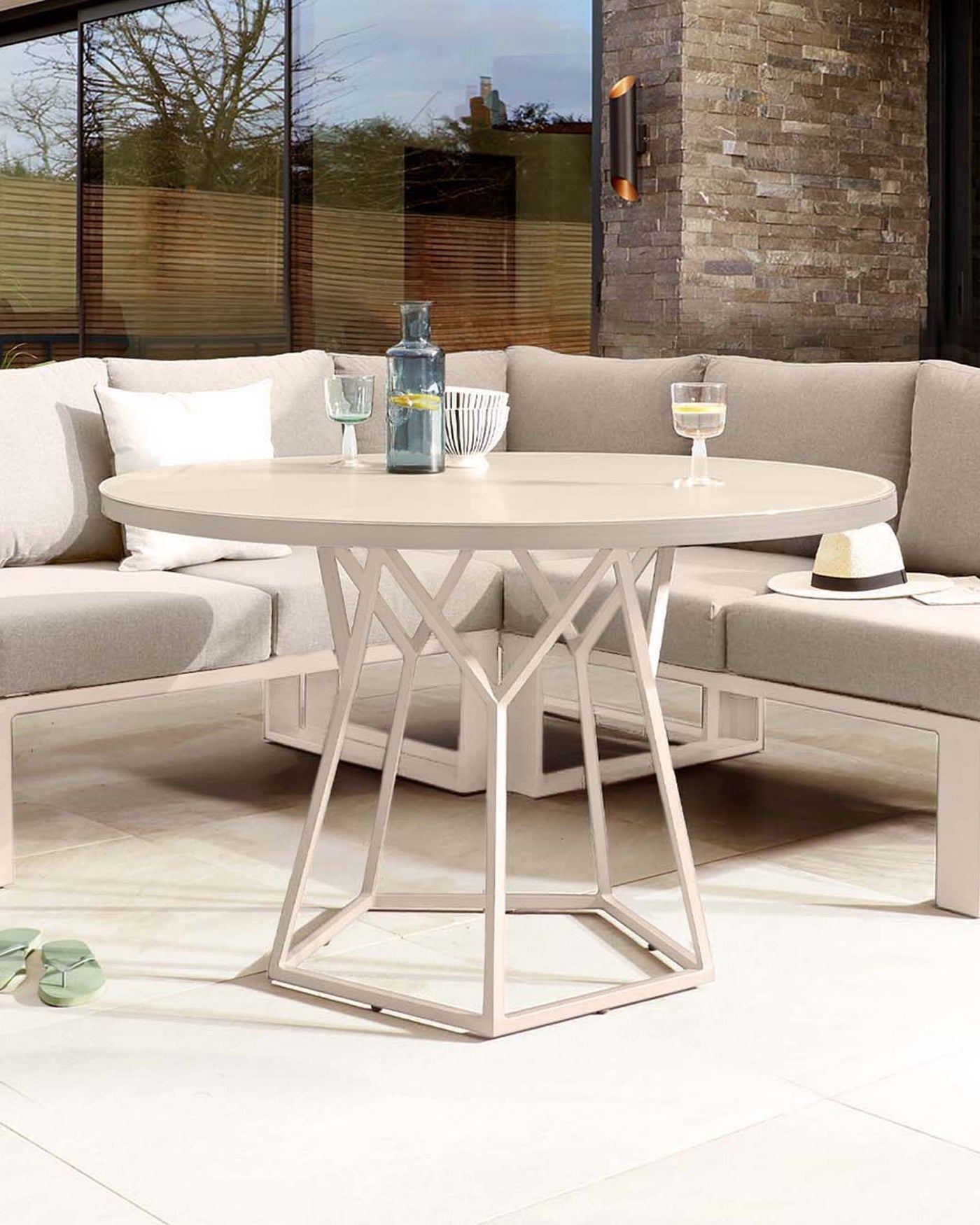 Modern outdoor furniture set featuring a round beige table with a unique geometric base and a matching L-shaped sectional sofa with plush cushions and pillows, staged on an elegant patio.