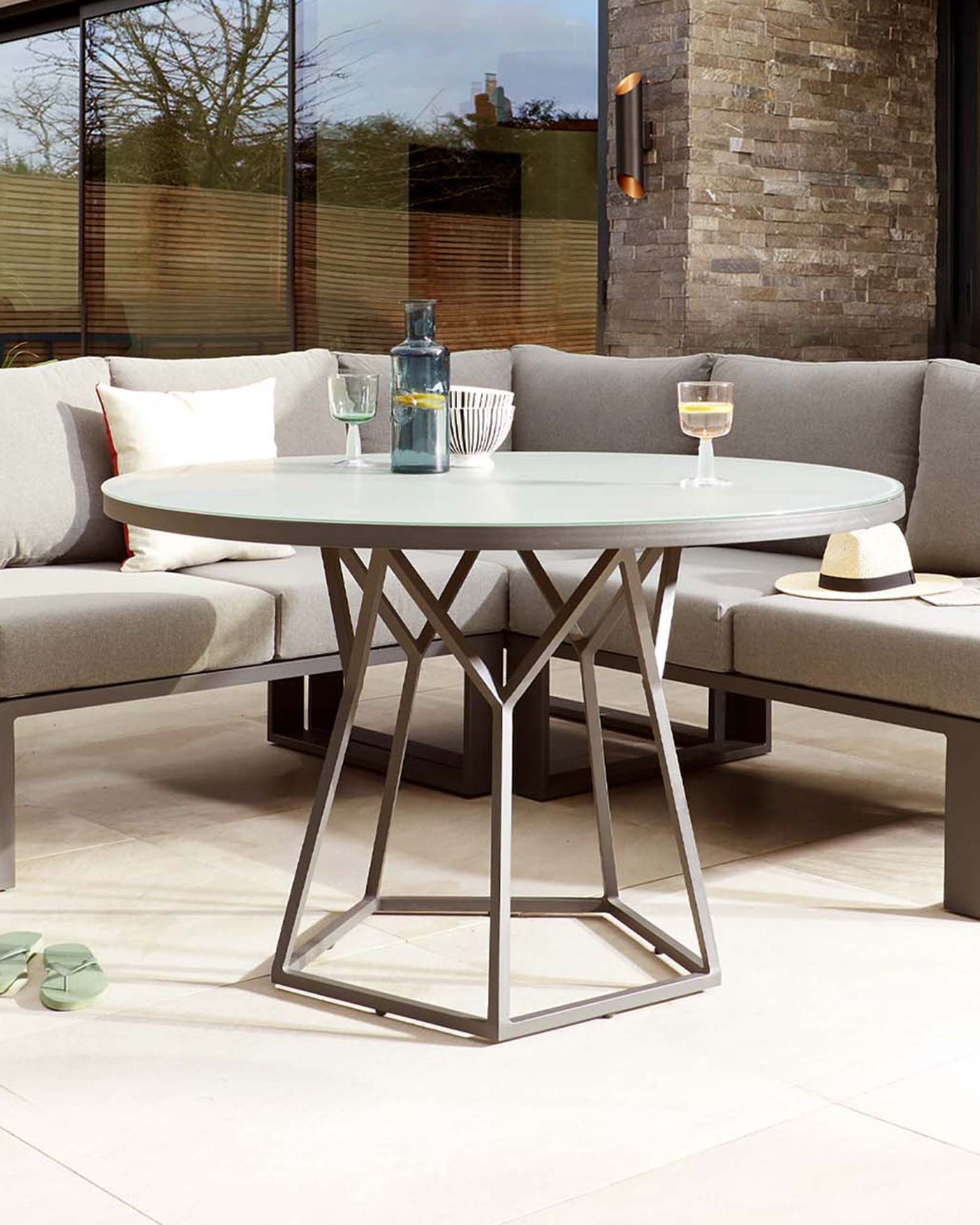 A modern outdoor round table with a frosted glass top and a geometric metallic base in a pentagonal prism shape, paired with a matching sectional outdoor sofa with light grey cushions.