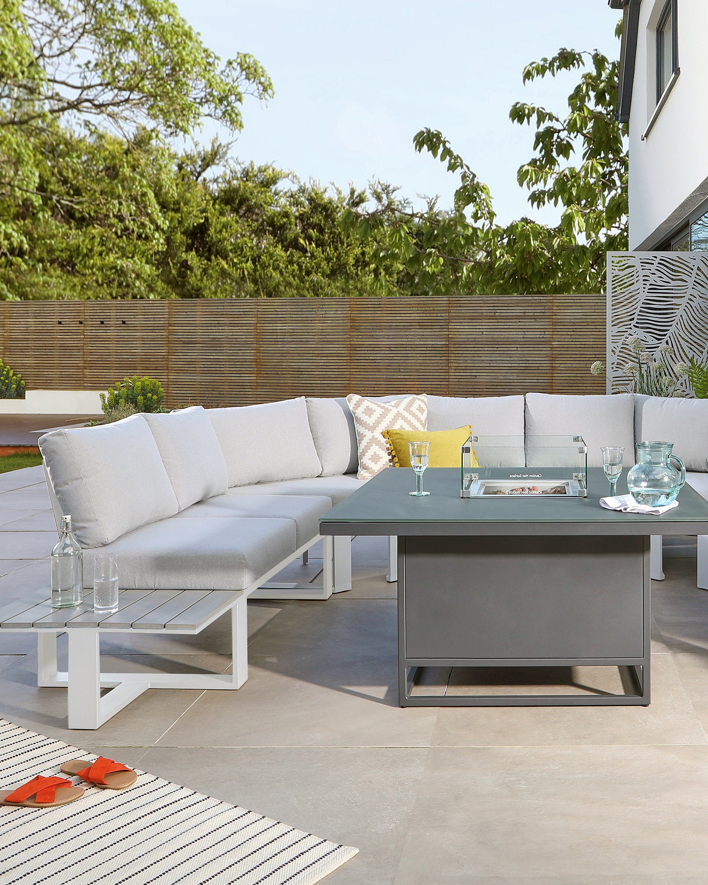 Modern outdoor furniture set featuring a sectional sofa with light grey cushions and a white frame, accompanied by a dark grey square coffee table with a glass top in a patio setting.