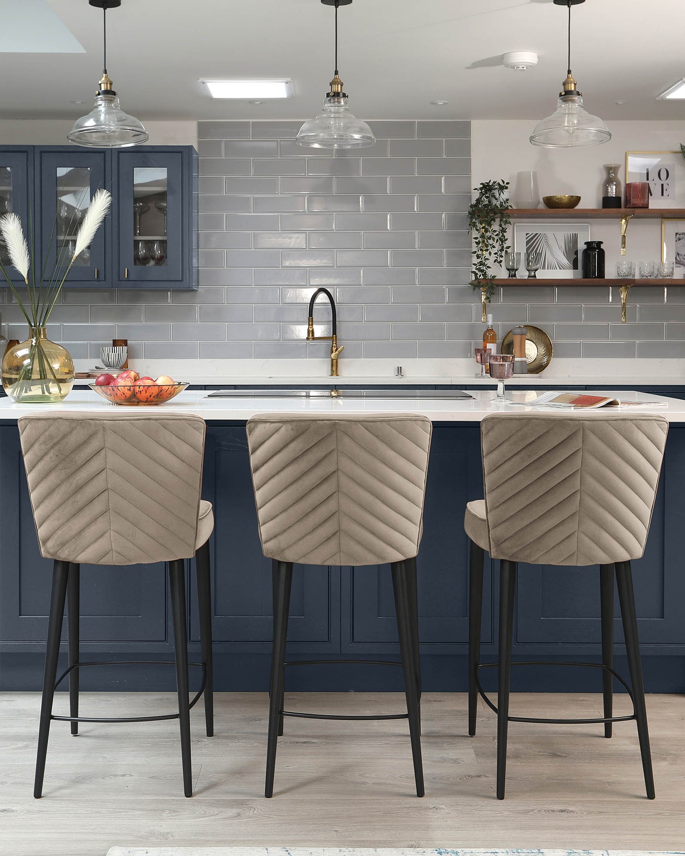 Three contemporary bar stools with upholstered seats in a light, textured fabric and chevron stitching. They feature curved backrests and slender black metal legs with footrests, positioned at a kitchen island.