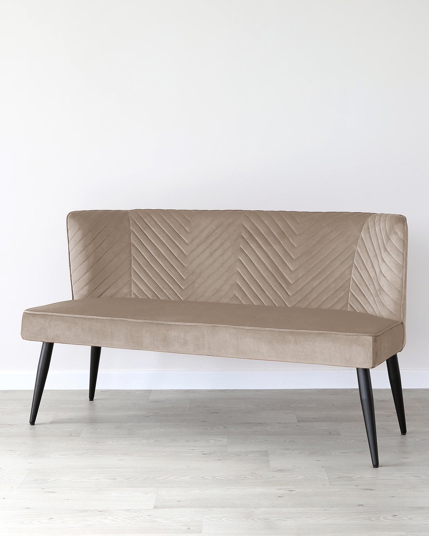 Modern upholstered bench with chevron-patterned backrest and seat in a light taupe fabric, complemented by four slanted black wooden legs. The bench is positioned against a plain white wall on a light wooden floor.