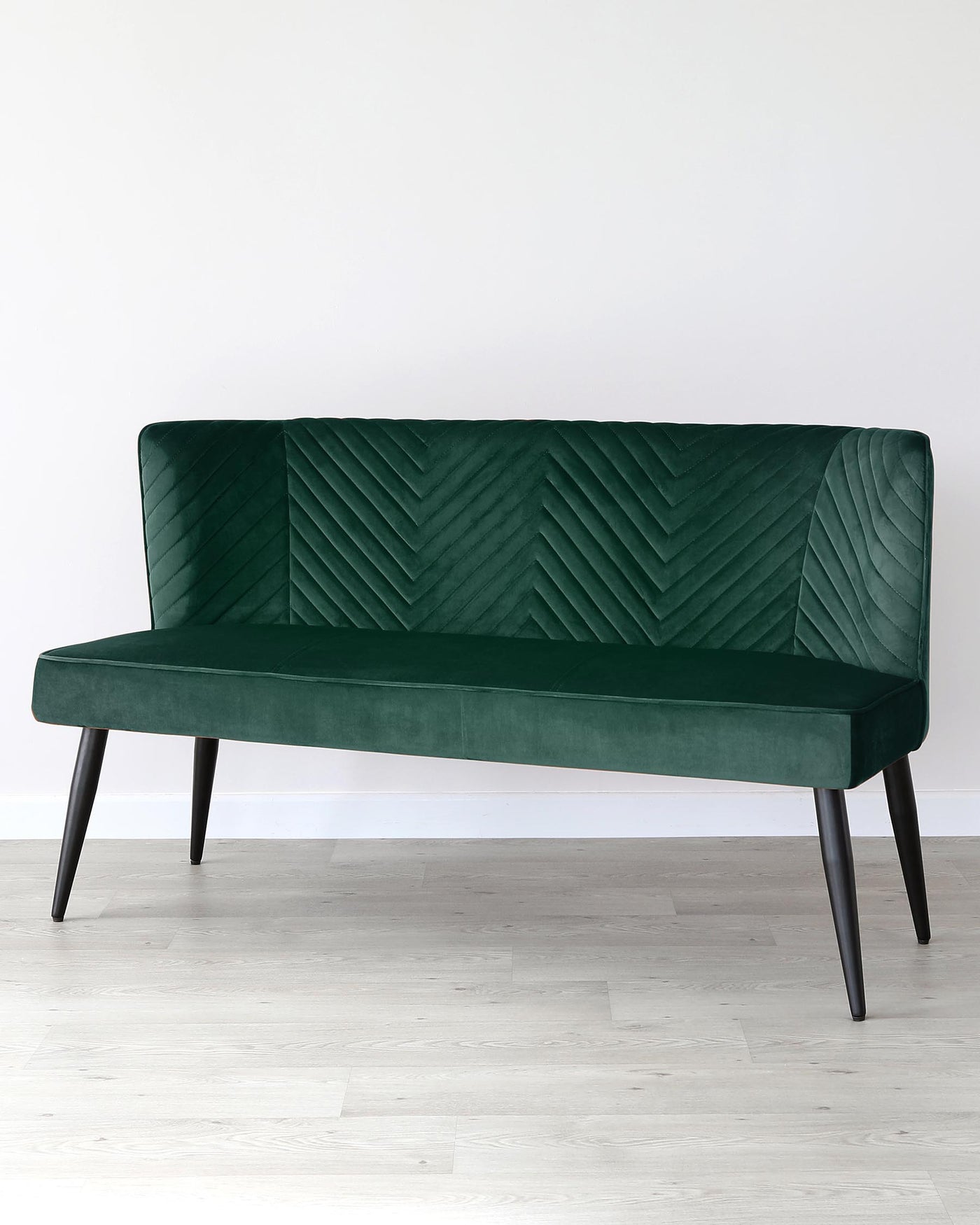 Elegant emerald green velvet settee with a high back, chevron-patterned tufting, and angled black wooden legs, presented against a plain white wall and light wooden flooring.