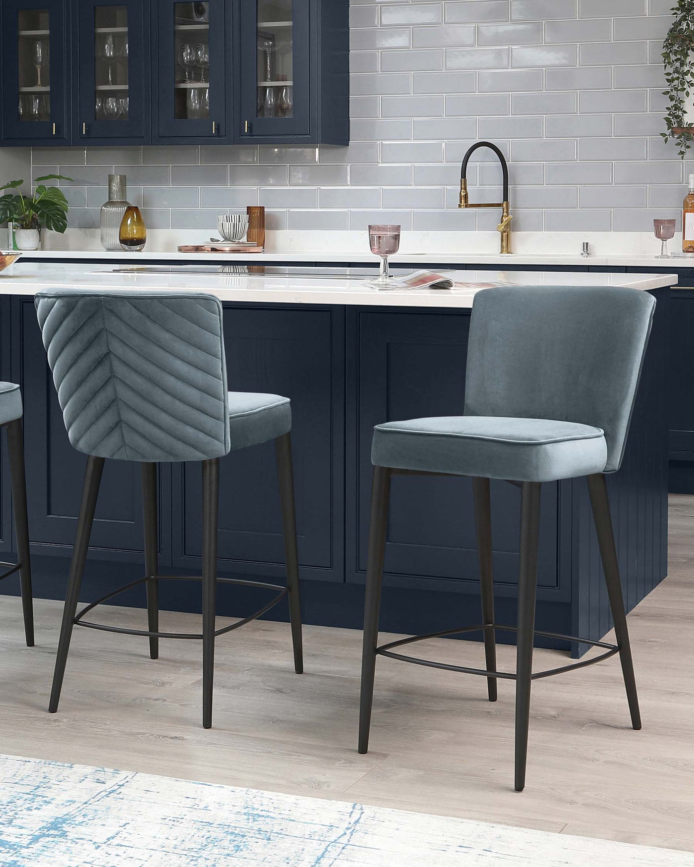 Elegant modern bar stools with sleek metal frames and luxurious quilted velvet upholstery in a soft grey tone, set against a kitchen island with a deep blue finish.