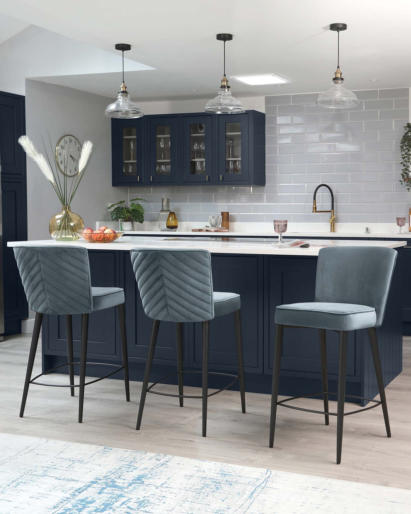 Three elegant bar stools with plush, channel-tufted velvet upholstery in a soft grey colour. Each stool has a high backrest and rests on slender, black metal legs with footrest bars, complementing a modern kitchen island setting.