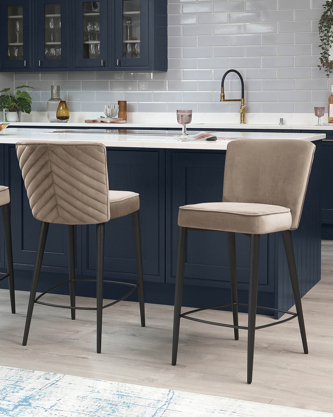 Two contemporary upholstered bar stools with channel tufting on the backrests, featuring a neutral beige fabric and slender black metal legs with footrest bars.