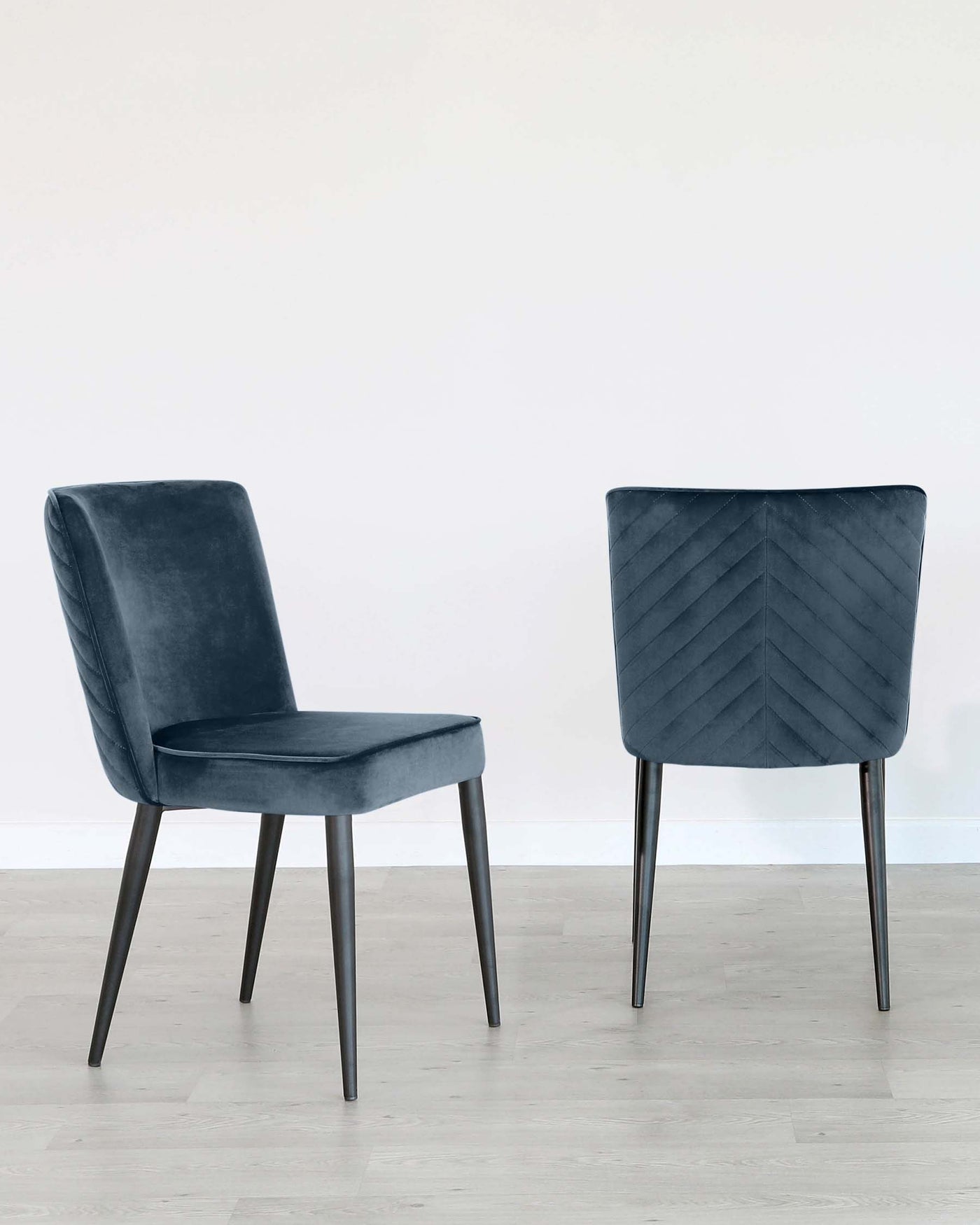 Two modern upholstered dining chairs with dark grey fabric and black metal legs, one featuring a smooth front and the other with a diagonally stitched backrest, standing on a light hardwood floor against a white wall.