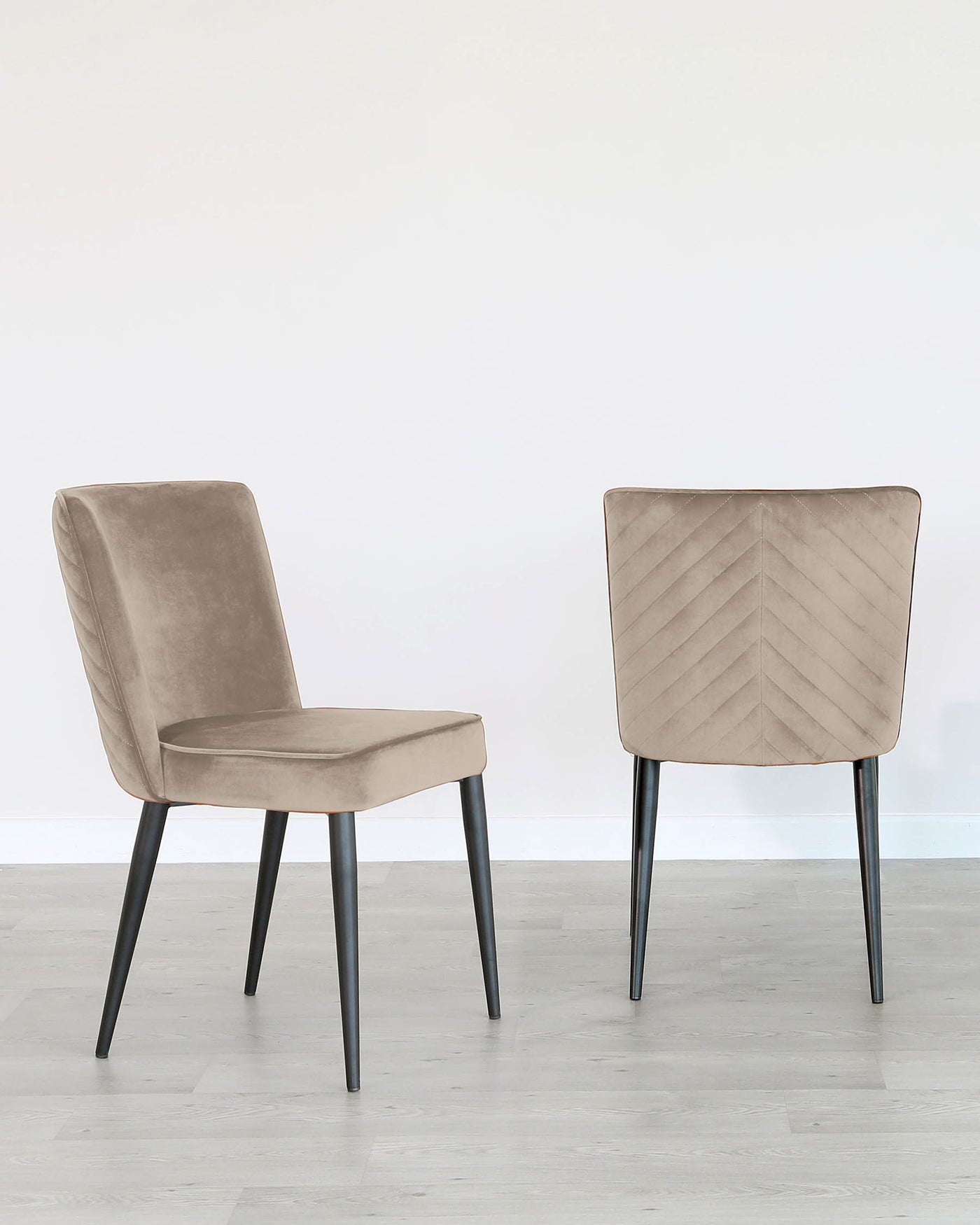 Two contemporary dining chairs with a minimalist design, featuring beige velvet upholstery and angular black metal legs. The chair visible from the front has a straight back, whereas the chair shown from the rear exhibits a distinctive chevron pattern stitching on the backrest.