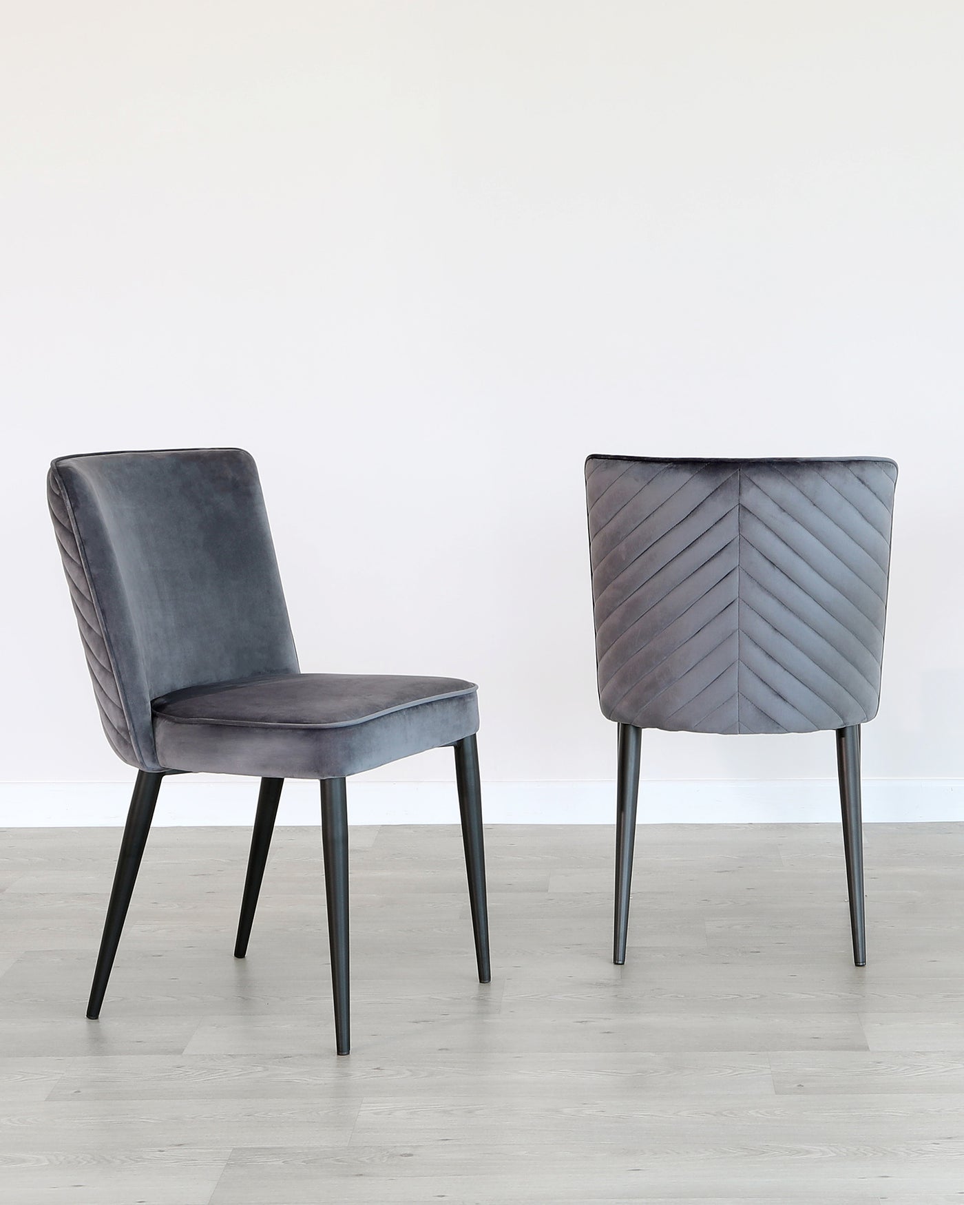 A pair of contemporary dining chairs with a luxurious grey velvet upholstery. The chairs feature a simple yet elegant design with a quilted backrest, displaying a chevron pattern, and sleek black metal legs. The chairs are positioned on a light wooden floor against a white wall, presenting a modern minimalist aesthetic.