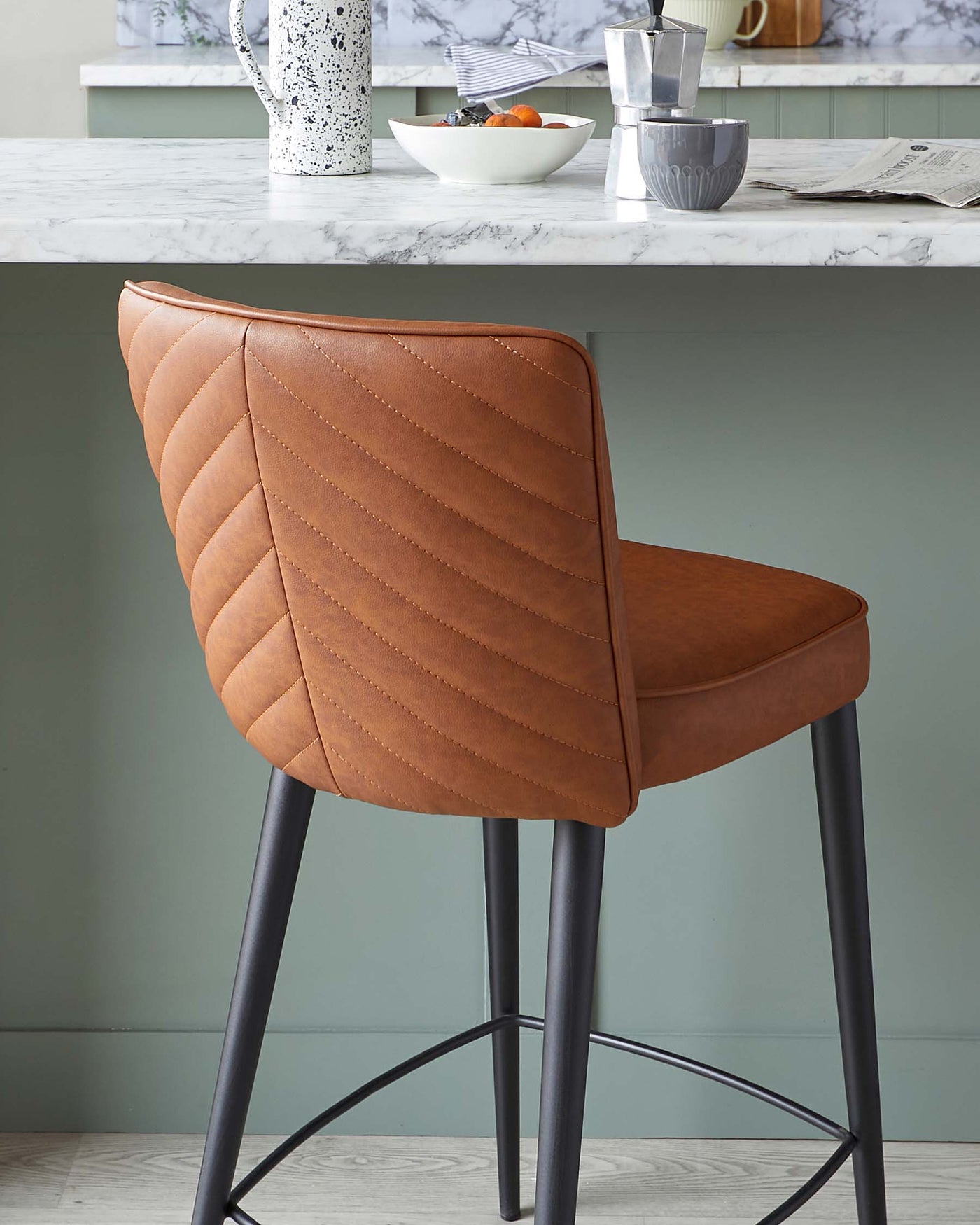Elegant modern bar stool with a quilted, caramel-coloured faux leather seat and a minimalist black metal frame, displayed in front of a kitchen countertop.