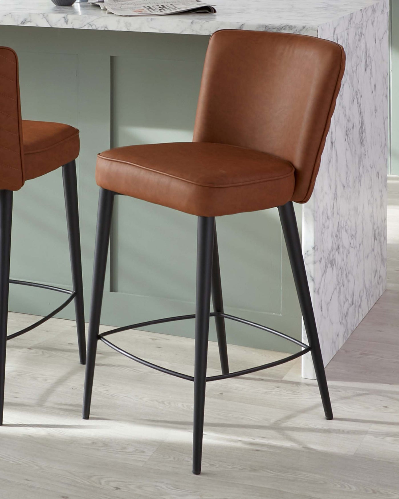 Modern brown faux leather bar stool with a padded back and seat with decorative stitching, anchored by a slender black metal frame with footrests. The stool complements a contemporary marble-patterned bar counter.