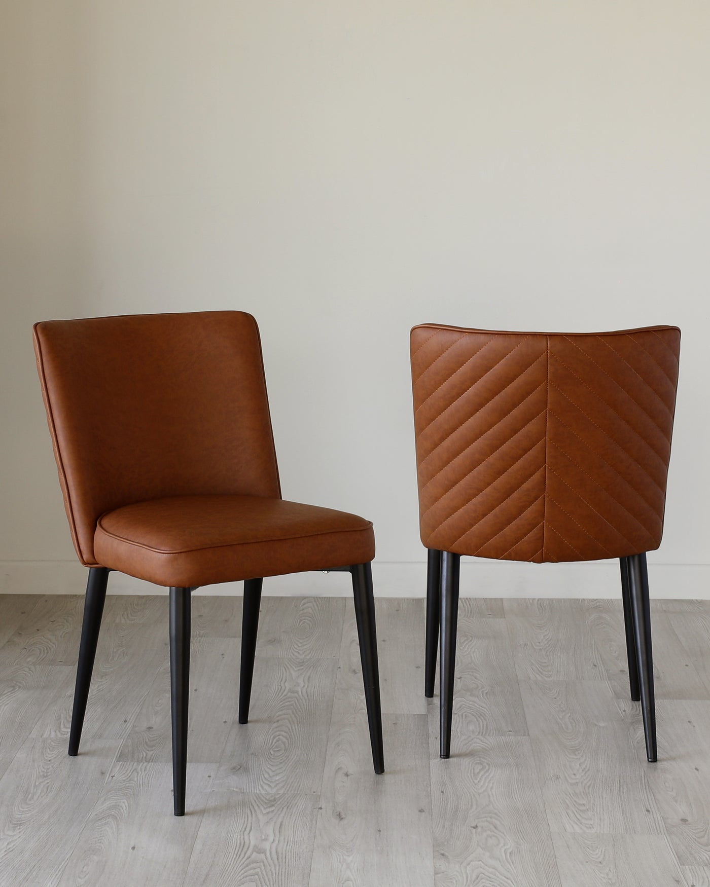 Two modern dining chairs with caramel brown faux leather upholstery and black metal legs, one chair showing the front profile with a smooth finish, and the other chair turned around displaying a diamond quilted backrest design.