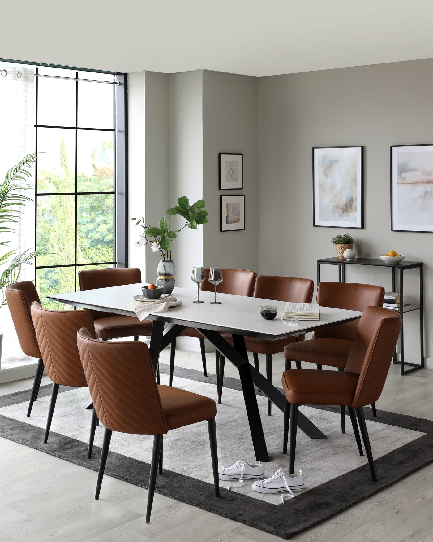 Modern dining room featuring a rectangular white marble-top table with black angular legs, surrounded by six caramel-brown upholstered dining chairs with quilted backrests and black metal legs. A minimalist black side table with books, a bowl, and decorative items is placed against the wall. The setup is on a grey area rug over light hardwood flooring.