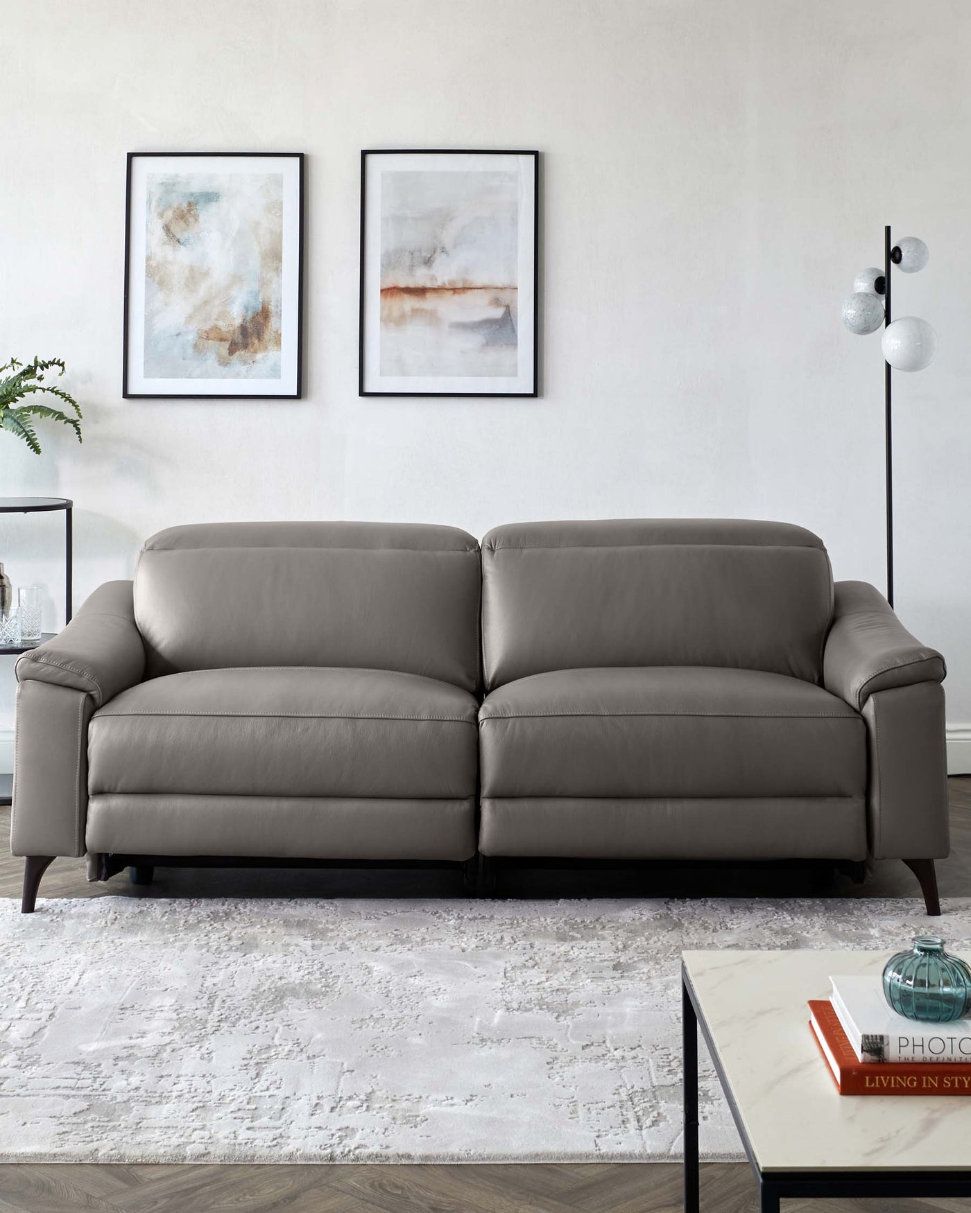 Modern grey leather sofa with clean lines and dark wooden legs, positioned on a textured off-white area rug, accompanied by a minimalist black metal floor lamp with glass globes, beside a sleek white marble-top coffee table with books and a decorative glass vase.