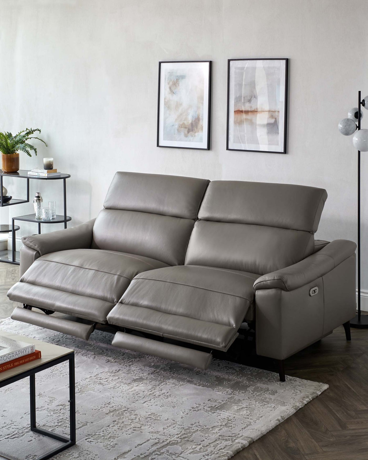 Modern grey leather sectional sofa with adjustable headrests and extended footrest on one end. Contemporary black metal and glass side table, along with a minimalist black frame coffee table with a glass top, on a textured off-white area rug.