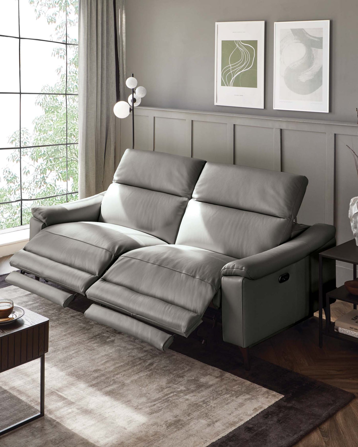 Modern three-seater leather recliner sofa in grey, featuring plush cushioning, with an adjustable backrest and footrest for comfort. A contemporary side table with a black metal frame and wooden top, along with an elegant floor lamp and grey-toned area rug, complements the chic and cosy living space setting.