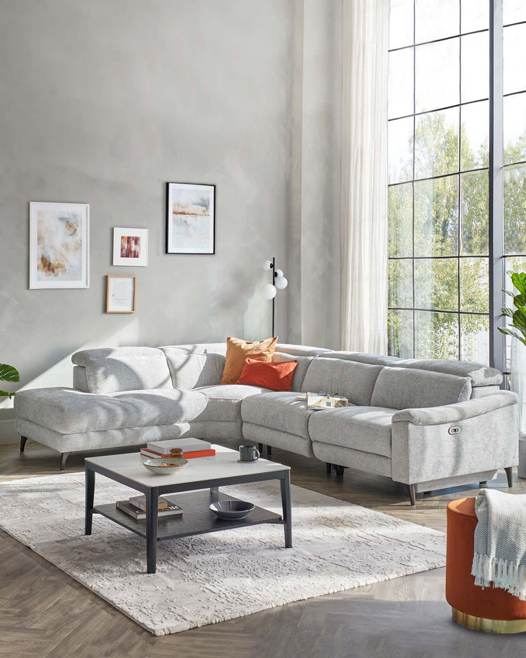Elegant living room set featuring a light grey sectional sofa with modern design, sleek lines, and tufted upholstery. A rectangular black and white coffee table with a lower shelf centres the area, complemented by a soft white area rug. An orange cylindrical stool with a gold base provides a pop of colour and an additional seating or decor option.