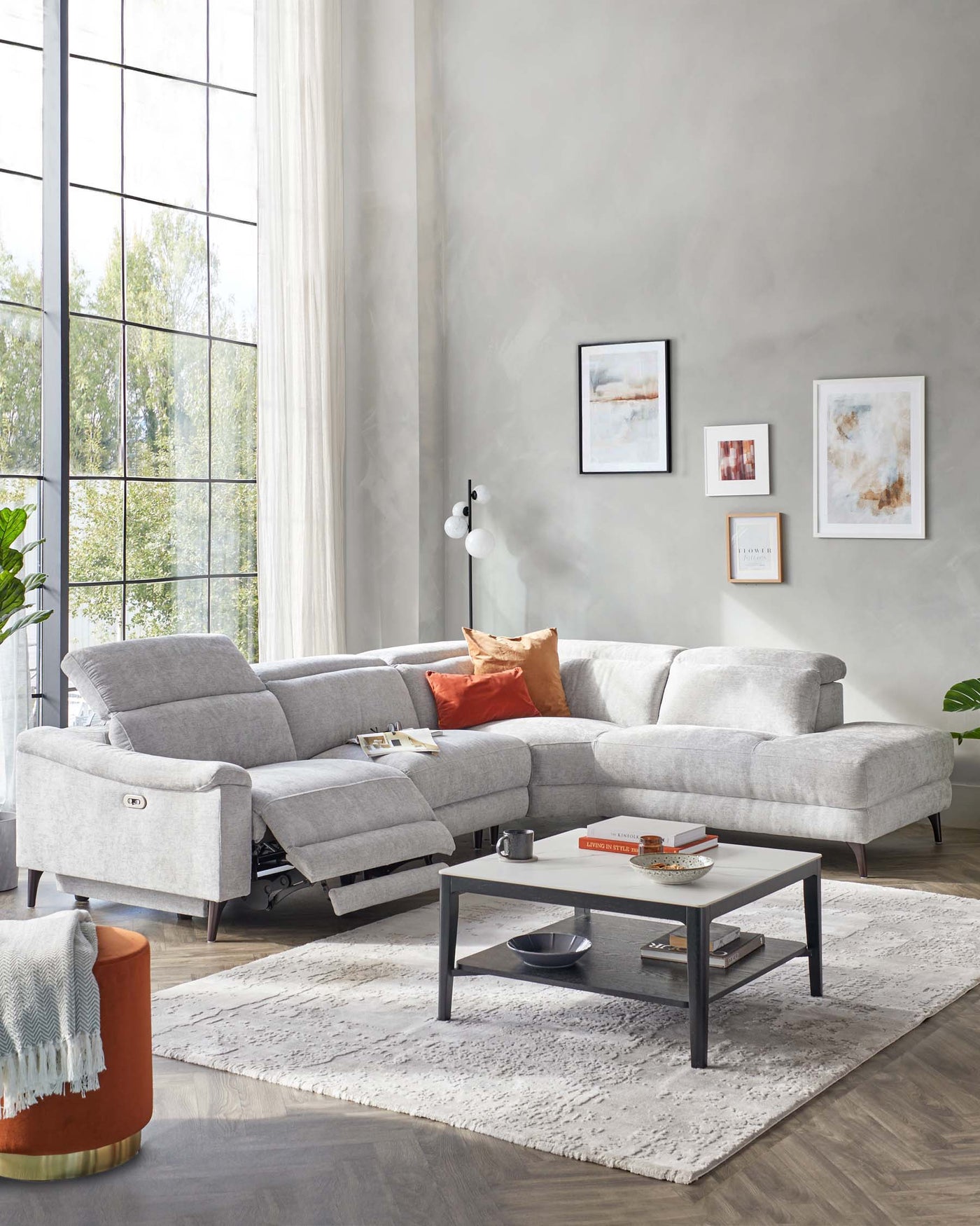 A modern living room featuring a light grey, L-shaped sectional sofa with plush cushions and contemporary metal legs. In the centre, a sleek, black rectangular coffee table with a glass top and shelf underneath sits on a textured off-white area rug. A round, burnt-orange side table with a gold-coloured base adds a pop of colour and complements the throw pillows on the couch.