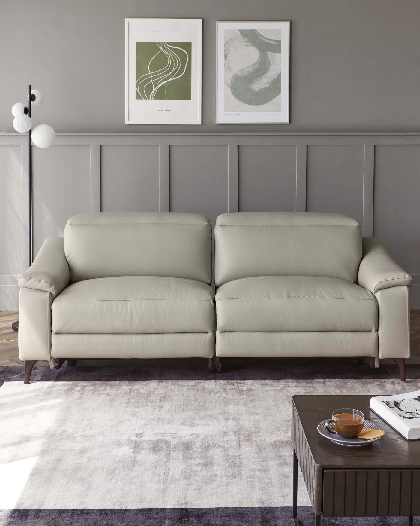 Elegant beige upholstered three-seater sofa with cushioned arms and dark wooden legs, paired with a dark wooden coffee table featuring a glass teapot and cup on top, set on a textured grey area rug.