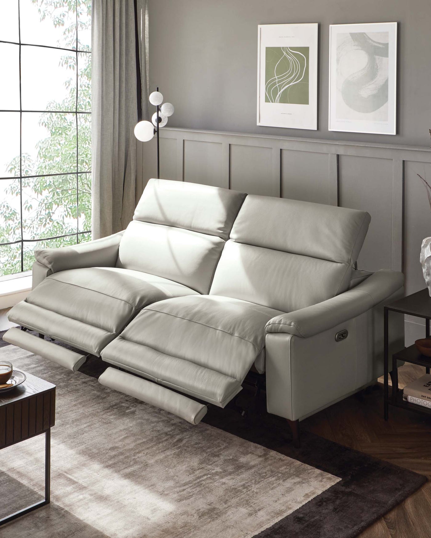 A modern light grey leather reclining sofa with padded armrests and headrest, set in a contemporary living room with a rectangular wooden side table, grey-toned area rug, and white spherical floor lamp. Two framed abstract wall art pieces hang above in a calm, neutral-toned space.