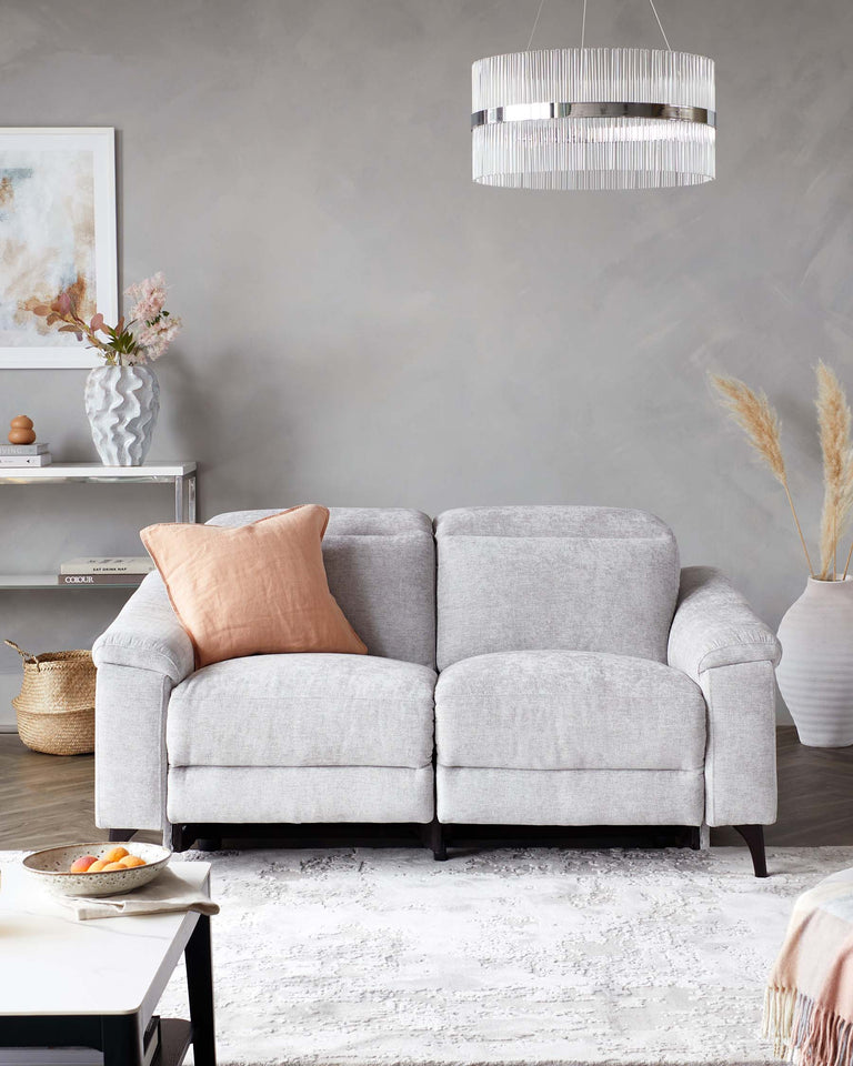 A contemporary light grey two-seater fabric sofa with plush cushioning and two round armrests, accentuated with a single blush pillow. The sofa stands on four dark wooden legs and is on a white textured area rug. In the background, a modern metallic pendant light hangs above, complementing the room's aesthetic. The scene includes a small white side table with minimalist decor and a larger console table with vases and ornamental items, implying a sophisticated and clean interior design.