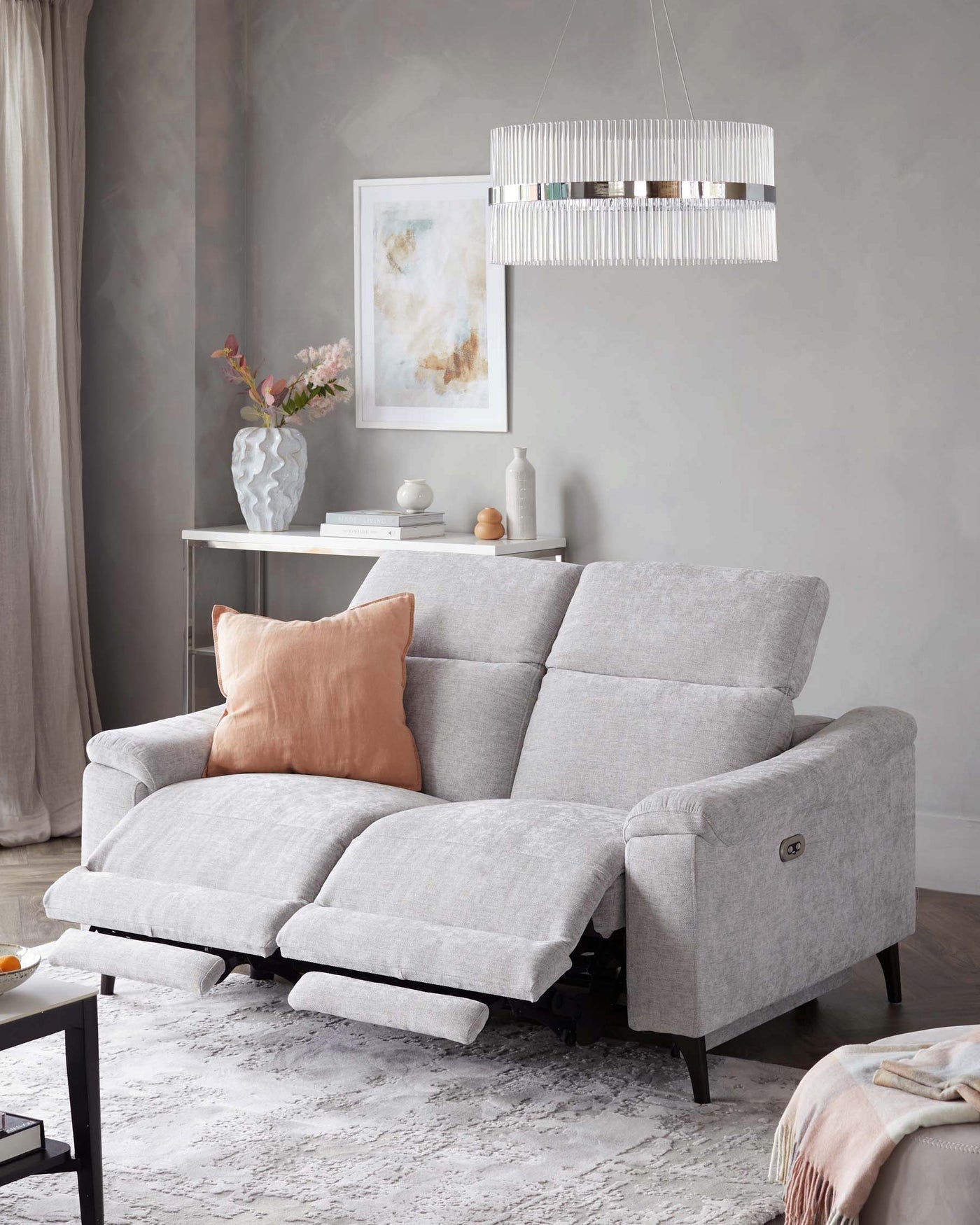 A light grey fabric sectional sofa with reclining footrests extended, complemented by pastel-coloured decorative pillows, set on a textured white area rug. Behind the sofa, a white console table decorates the room alongside a white vase with pink flowers, ceramic decor, and books, under a framed abstract art piece. Above the setting hangs a contemporary chandelier with vertical fringe elements.