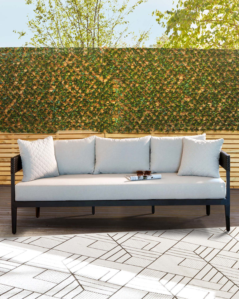 A modern outdoor sofa with a sleek wooden frame and light grey cushions, complemented by two patterned throw pillows, against a natural leafy backdrop. A small wooden tray with a pair of sunglasses rests on the sofa.