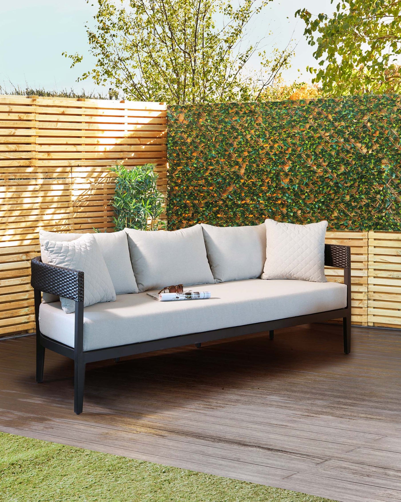 An outdoor sectional sofa with a modern design, featuring a black, minimalist frame and light grey cushions, complemented by a series of off-white accent pillows. The sofa is situated on a wooden deck surrounded by greenery.