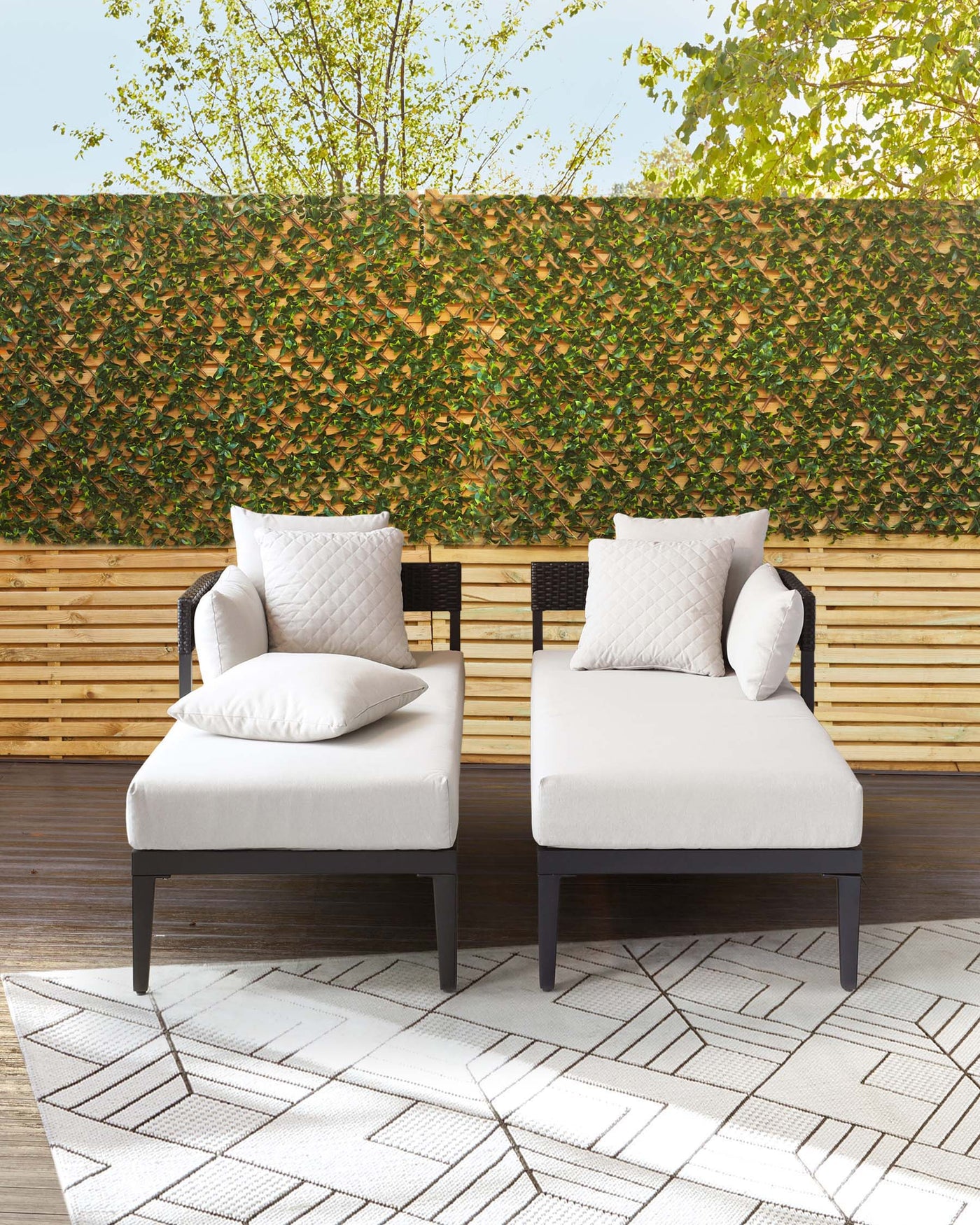 Modern outdoor furniture set featuring two armless, cushioned chairs with dark wooden frames and white upholstery, complemented by matching accent pillows on a geometric-patterned outdoor rug.