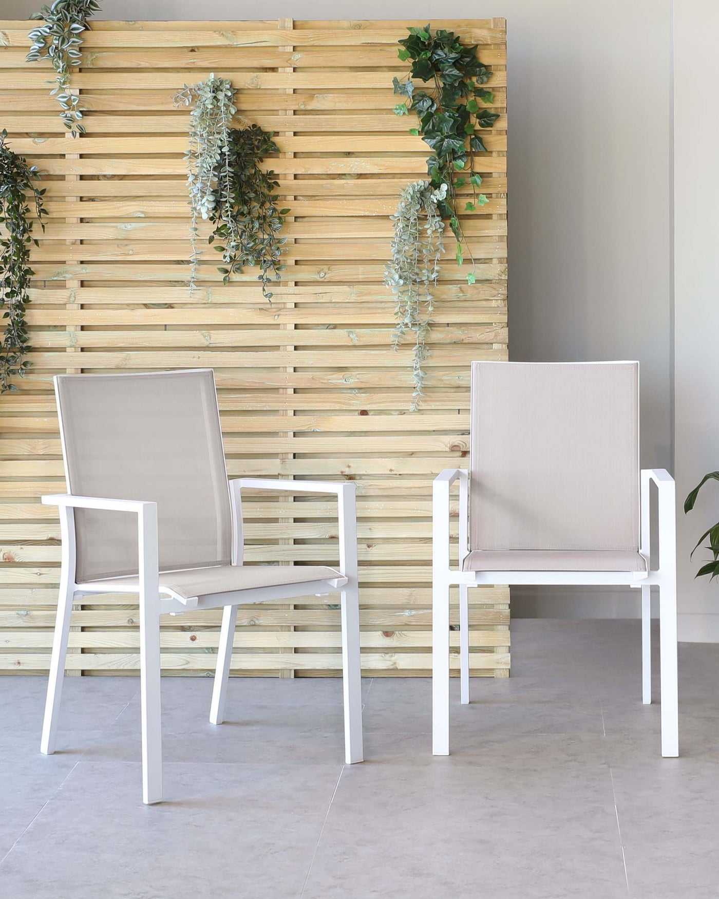 Two modern outdoor chairs with white metal frames and light taupe fabric seats and backs, set against a wooden slatted wall with hanging green plants.