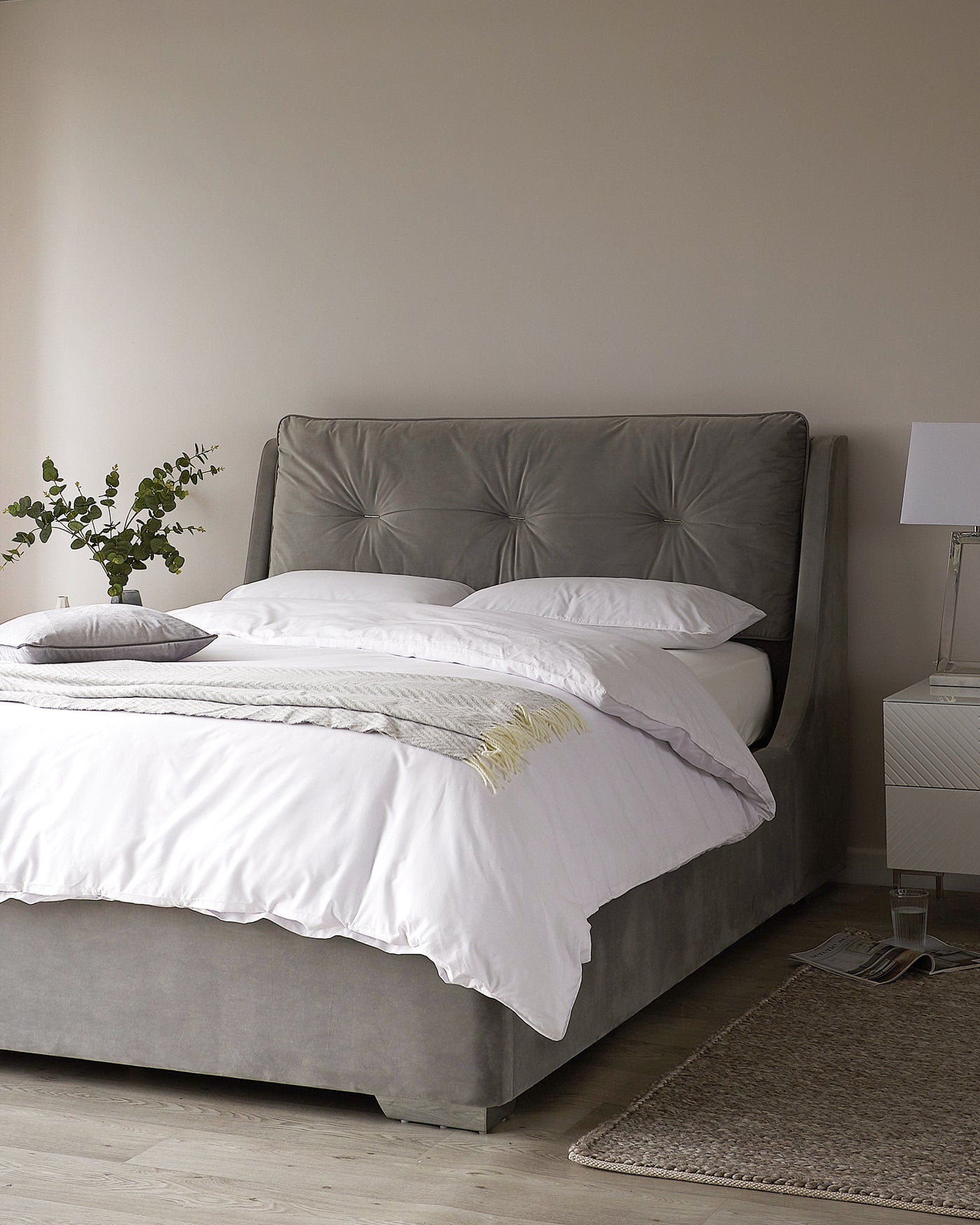 Elegant bedroom featuring a large, grey upholstered bed with a tufted headboard, white bedding, and a decorative throw pillow. A sleek white bedside table with a modern lamp is positioned to the right of the bed, while a textured grey area rug lies partially underneath. A green potted plant adds a touch of nature to the space.