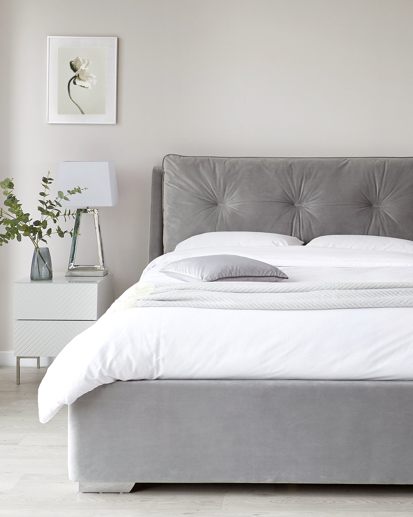 Elegant grey upholstered bed with a tall, tufted headboard and a clean-lined white nightstand with a chevron pattern. The bed is adorned with crisp white bed linens and fluffy pillows, and the nightstand features a contemporary white lamp with a clear base.