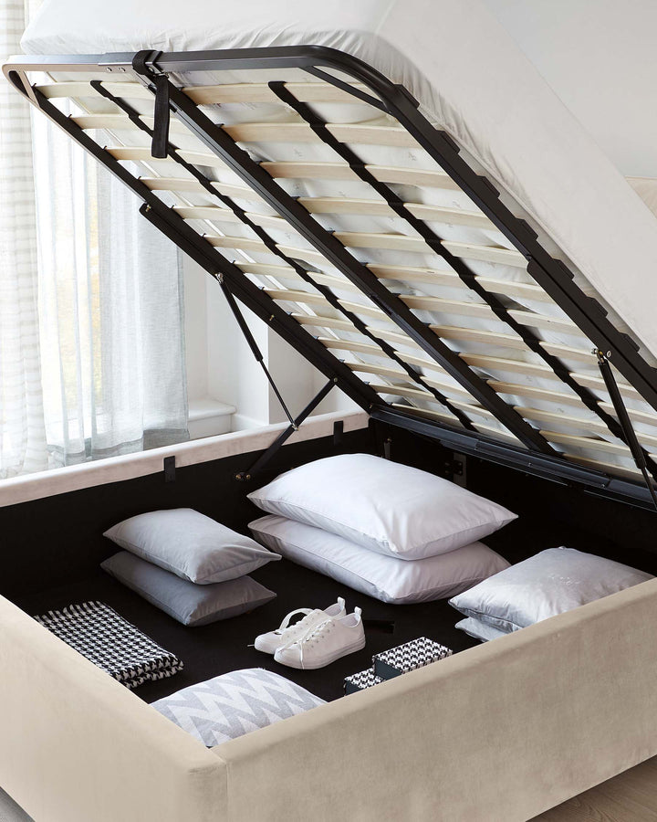 Beige upholstered storage bed with a lifted mattress platform revealing a spacious storage compartment containing pillows, blankets, and a pair of shoes. The bed features a gas-lift mechanism with black metal framing supporting wooden slats under the mattress.