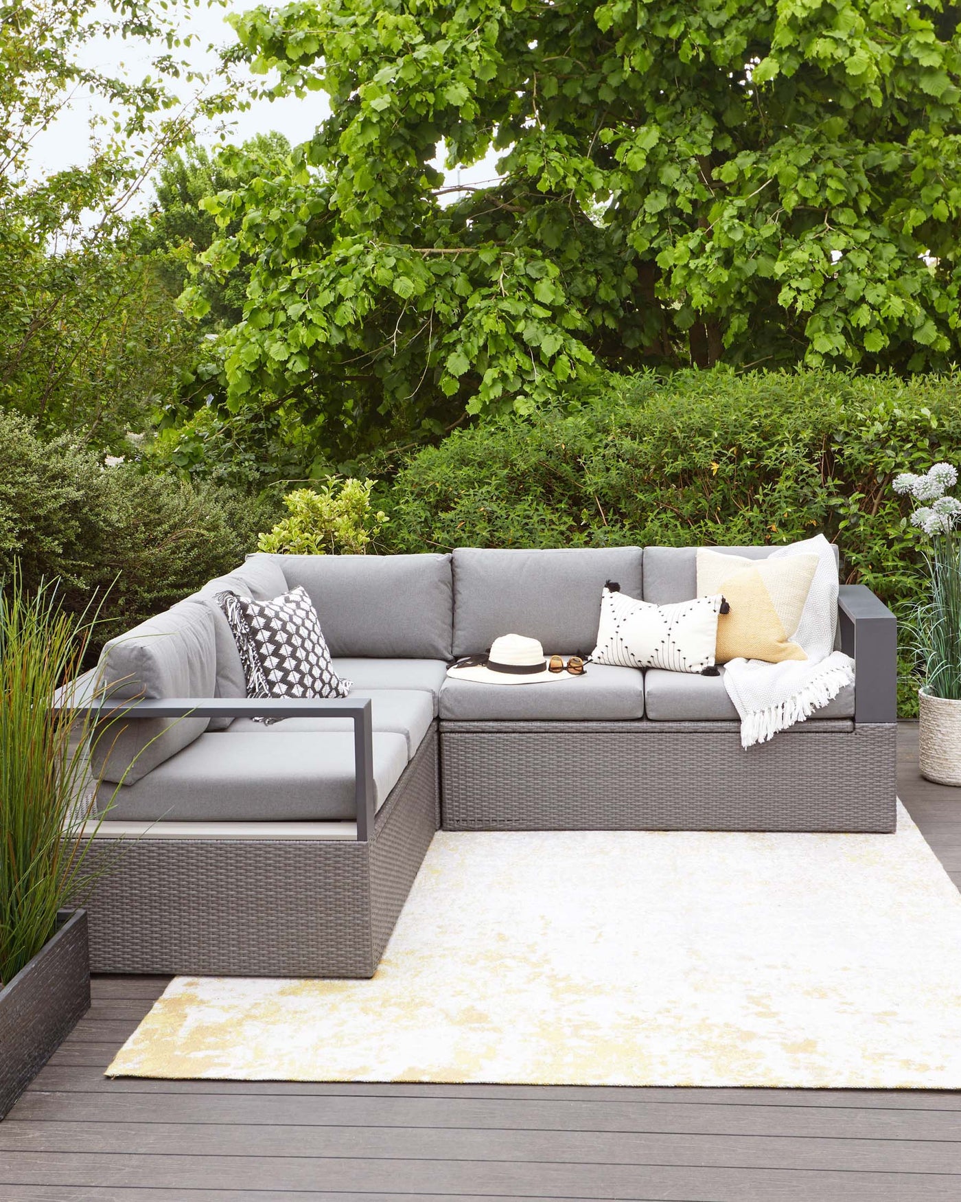 A modern outdoor sectional sofa with a chaise lounge in a woven grey resin wicker material, with thick light grey cushions. Coordinating decorative pillows in various patterns and a yellow accent are placed on the sofa. The furniture is situated on a cream and beige area rug, laid on a grey wooden decking. Set against a backdrop of lush greenery, the arrangement creates an inviting outdoor living space.