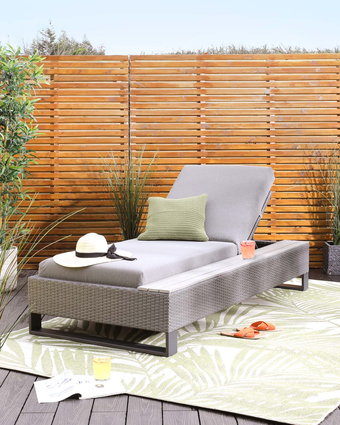 Contemporary outdoor furniture featuring a wicker chaise lounge with a grey cushion and pillow, accompanied by a matching wicker coffee table, set on a green patterned outdoor rug.