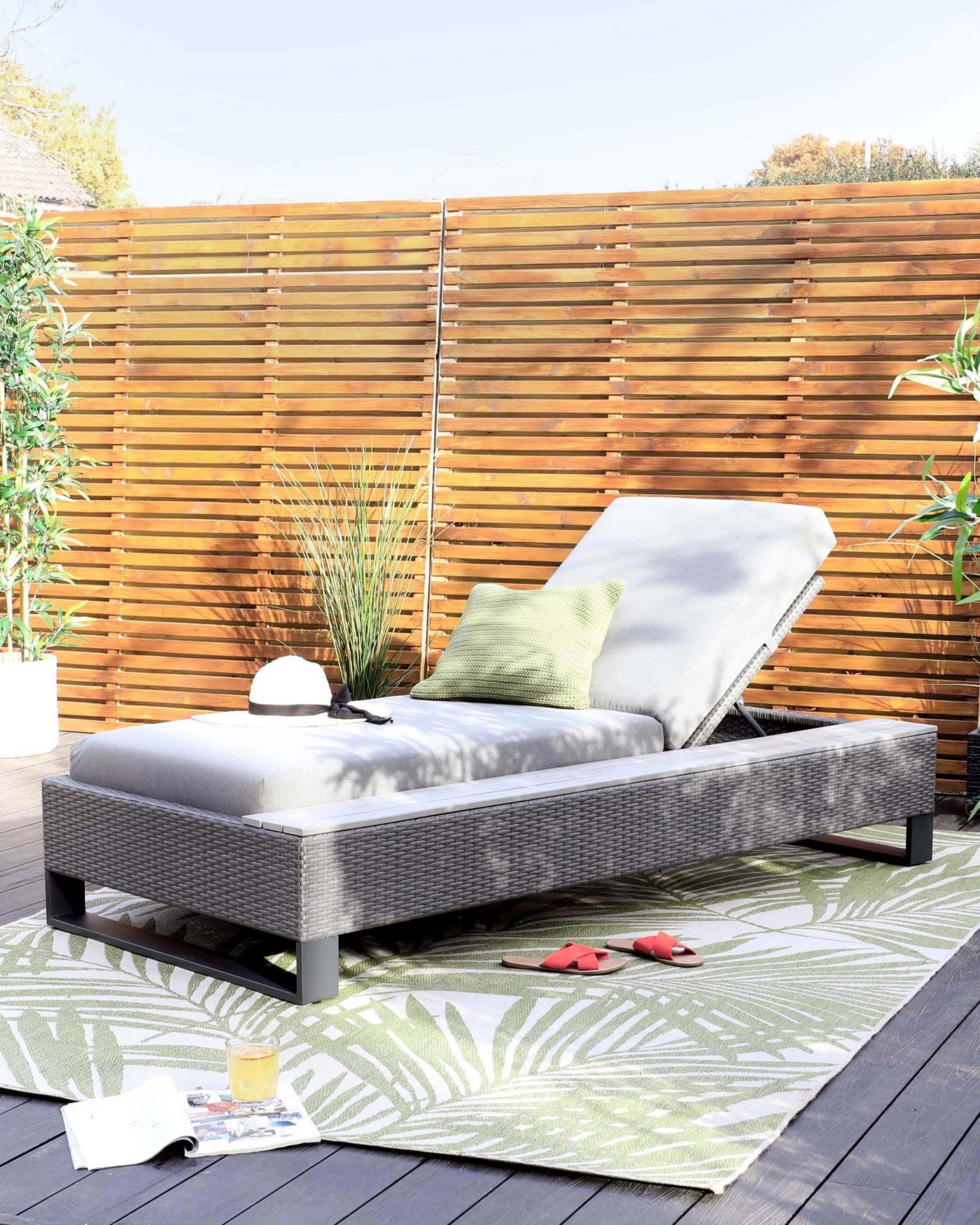 Outdoor chaise lounge with a grey wicker frame, adjustable backrest, and thick cushion atop a green and white patterned area rug on a wooden deck.
