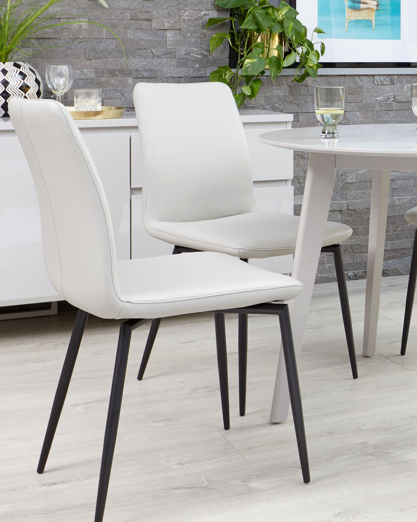 Modern minimalist furniture set including a white round dining table and sleek chairs with white upholstery and black metal legs, arranged on a light wood floor against a backdrop of a grey stone wall with decorative items and greenery.