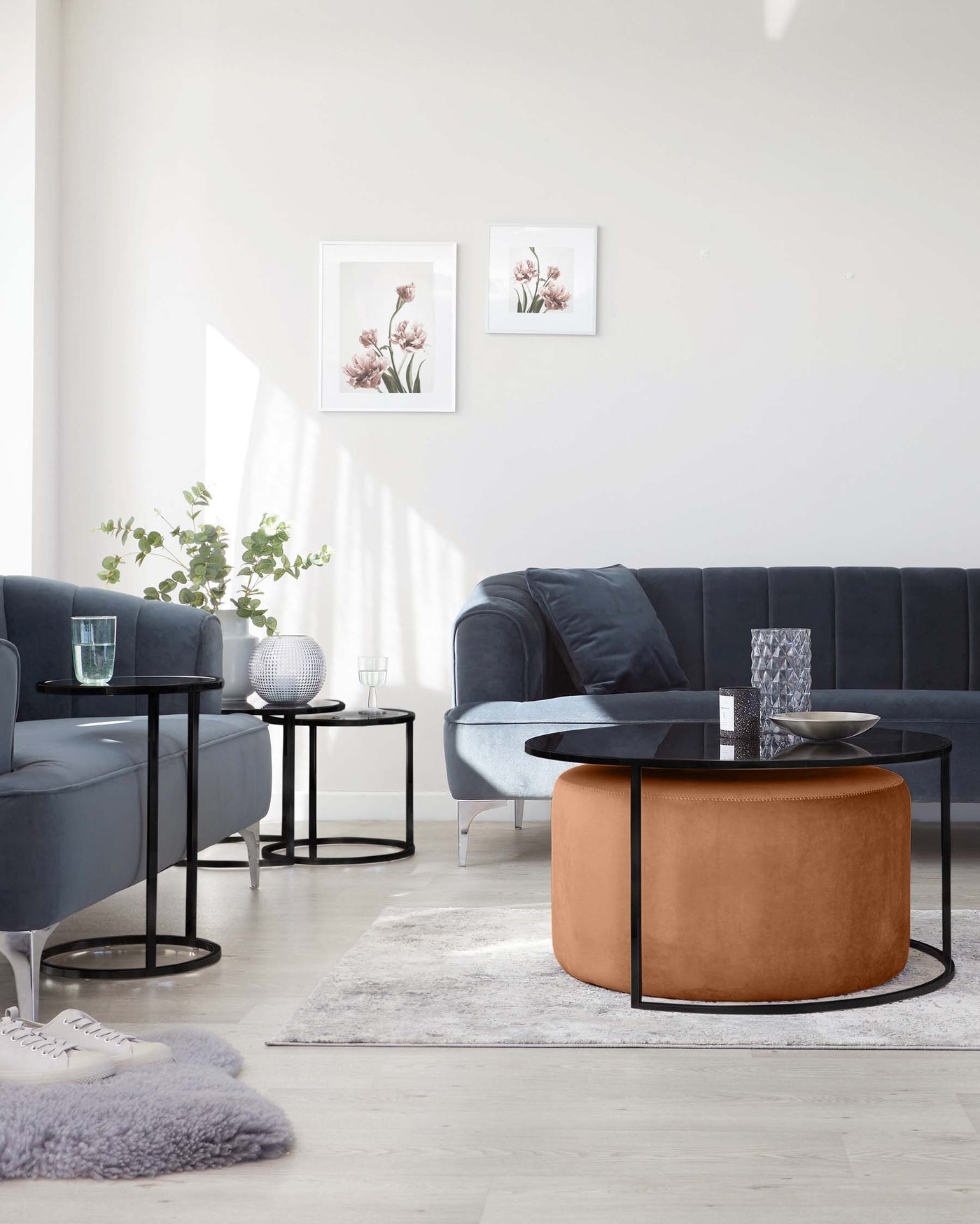 Modern living room showcasing a dark blue upholstered sofa with button detailing, flanked by two round black metal side tables with glass tops. In the centre stands a two-tiered round coffee table; the upper level is black with a glass surface, while the lower is a saddle tan leather pouf. A light grey area rug anchors the set against a simple backdrop.